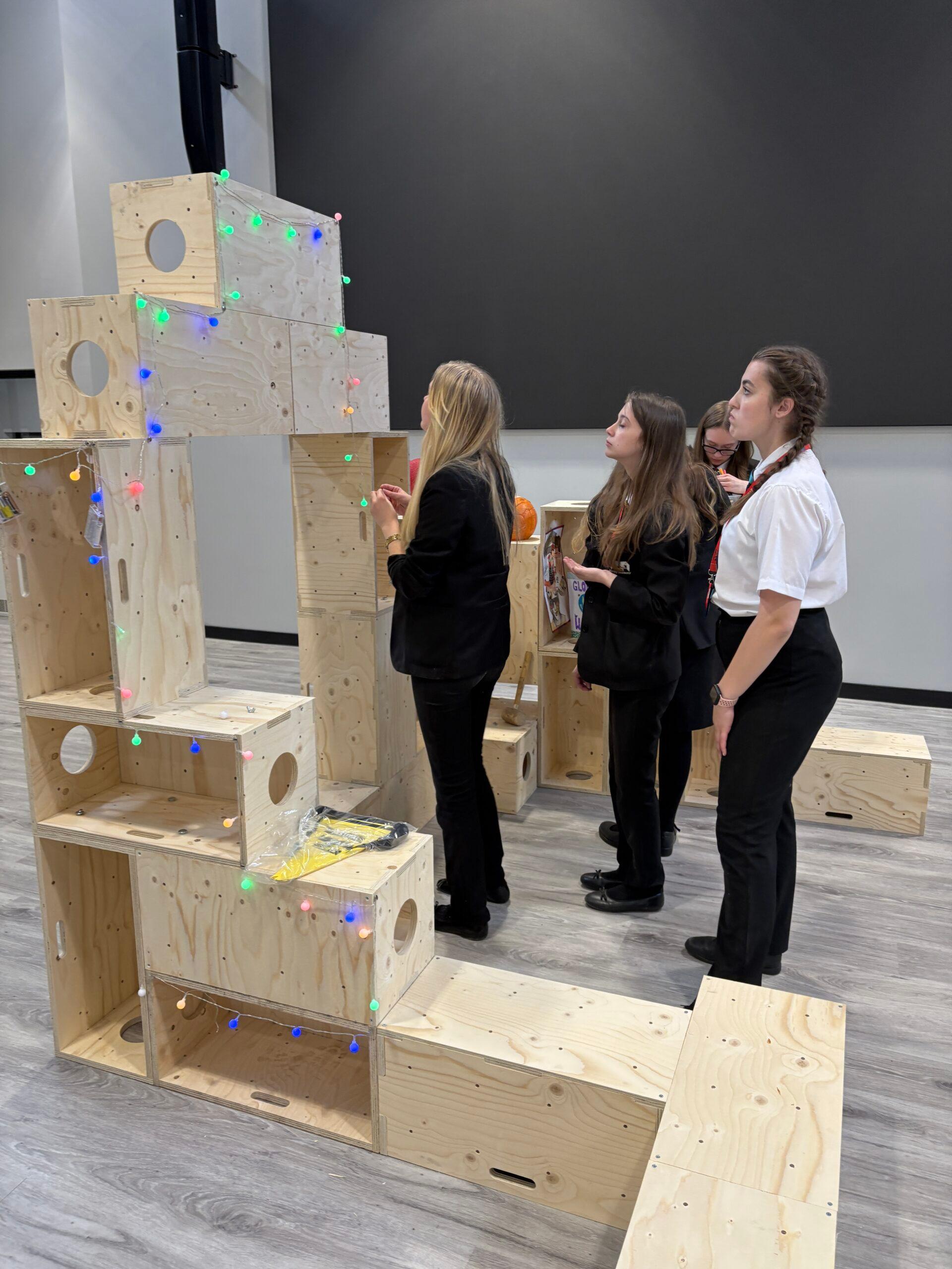 Four students in black uniforms interact with a large plywood block structure, decorated with colourful string lights, inside a modern indoor space featuring grey flooring and black walls.