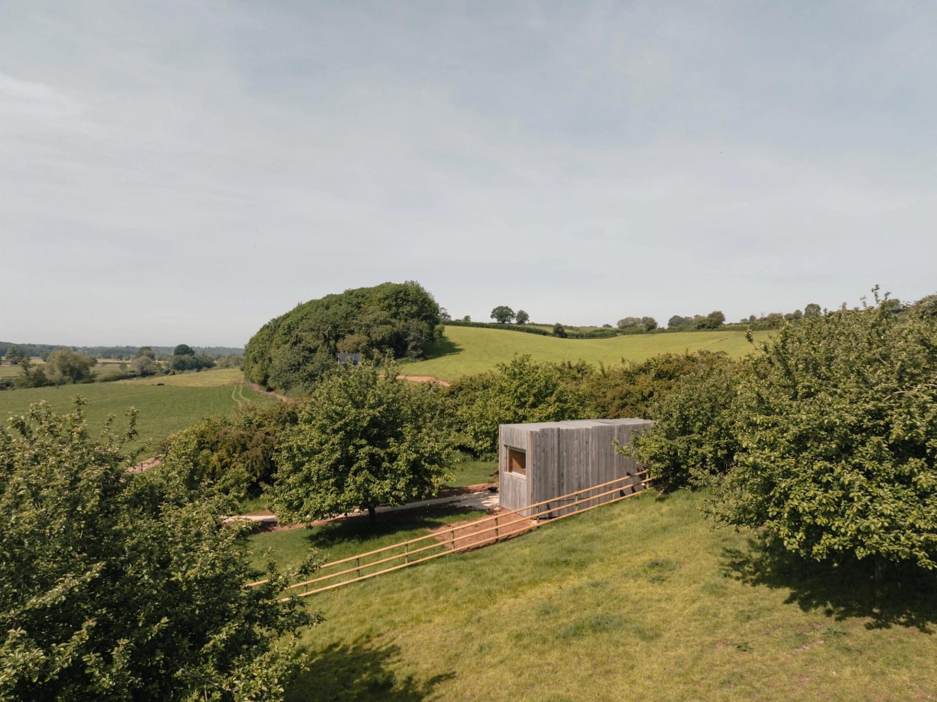 A small wooden cabin, featuring charming cabin furniture, sits among green trees and rolling hills under a clear sky, with a wooden fence and a dirt path leading towards the building.