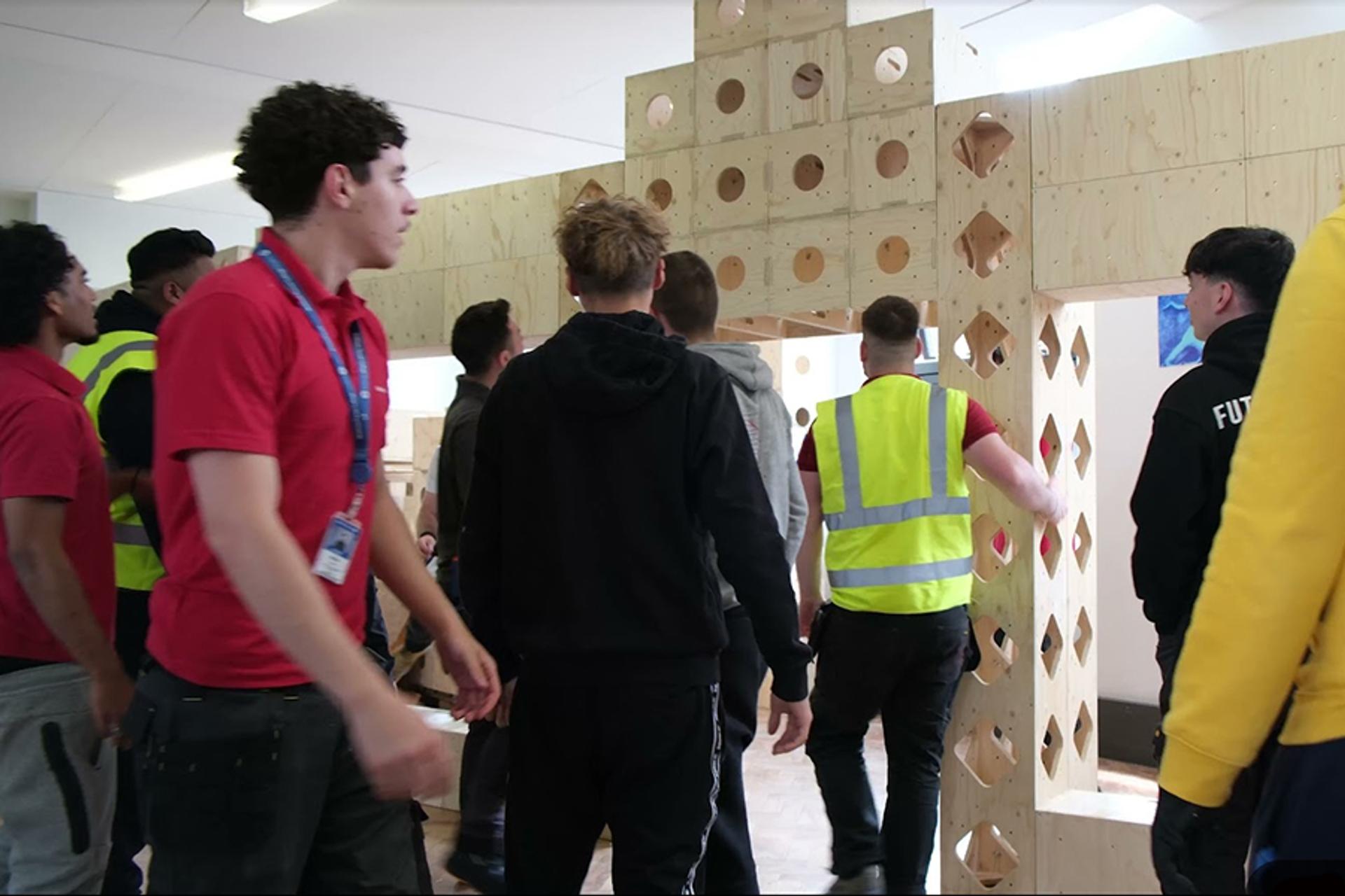 A group of people, some wearing high-visibility vests, stand and walk around a large sustainable birch plywood structure with geometric cut-outs inside a bright indoor space.