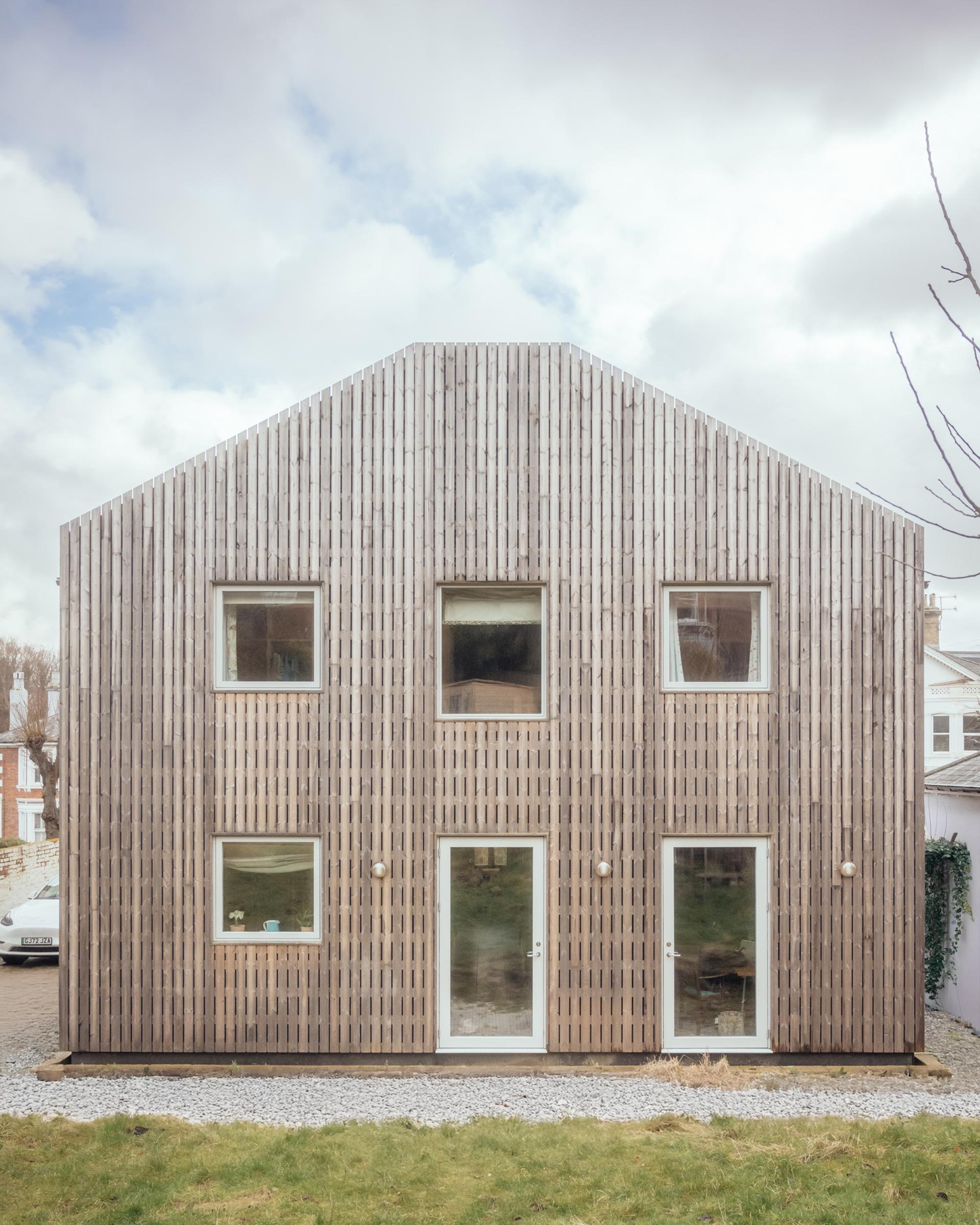 A two-storey house with a gabled roof and vertical timber cladding, featuring symmetrical windows and two glass doors on the ground floor.