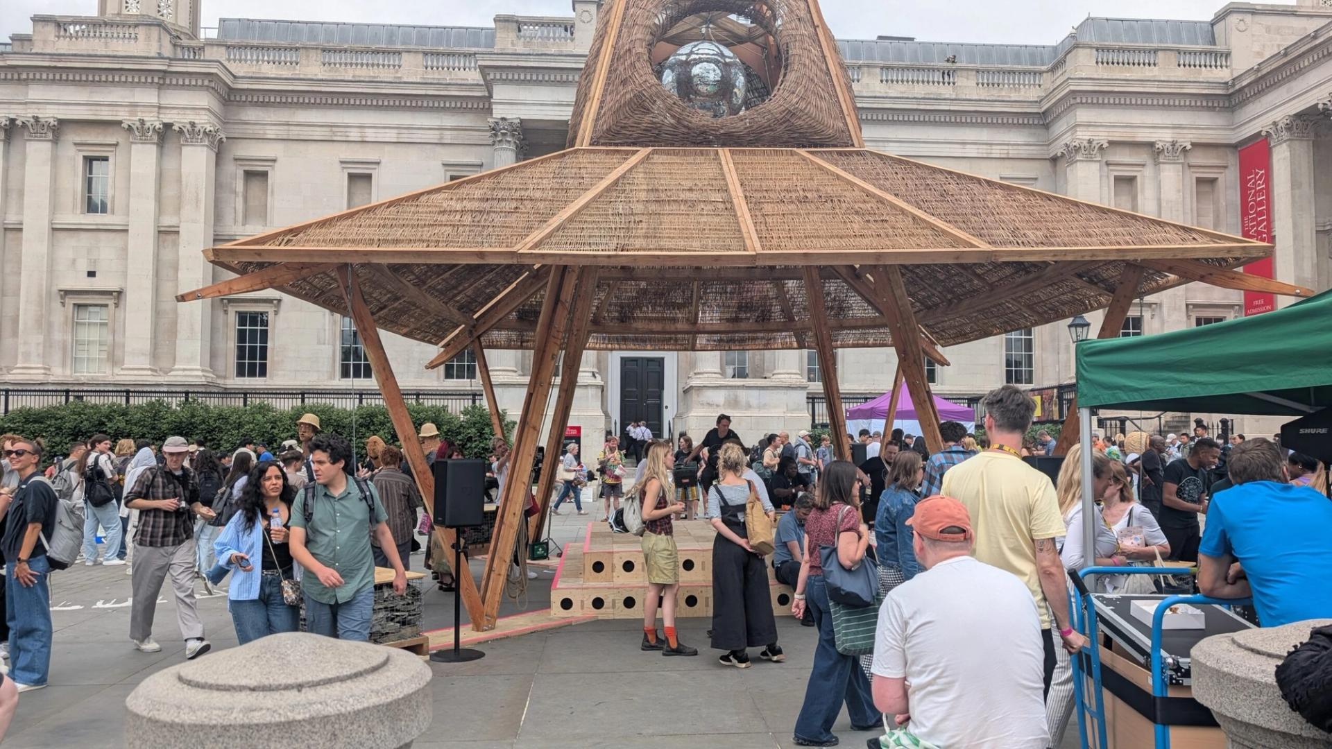 A crowd gathers around a large, wooden, pavilion-like art installation in an outdoor plaza near the National Gallery. People walk, talk, and interact with the structure as classical-style buildings frame this 2025 Triumph of Art.