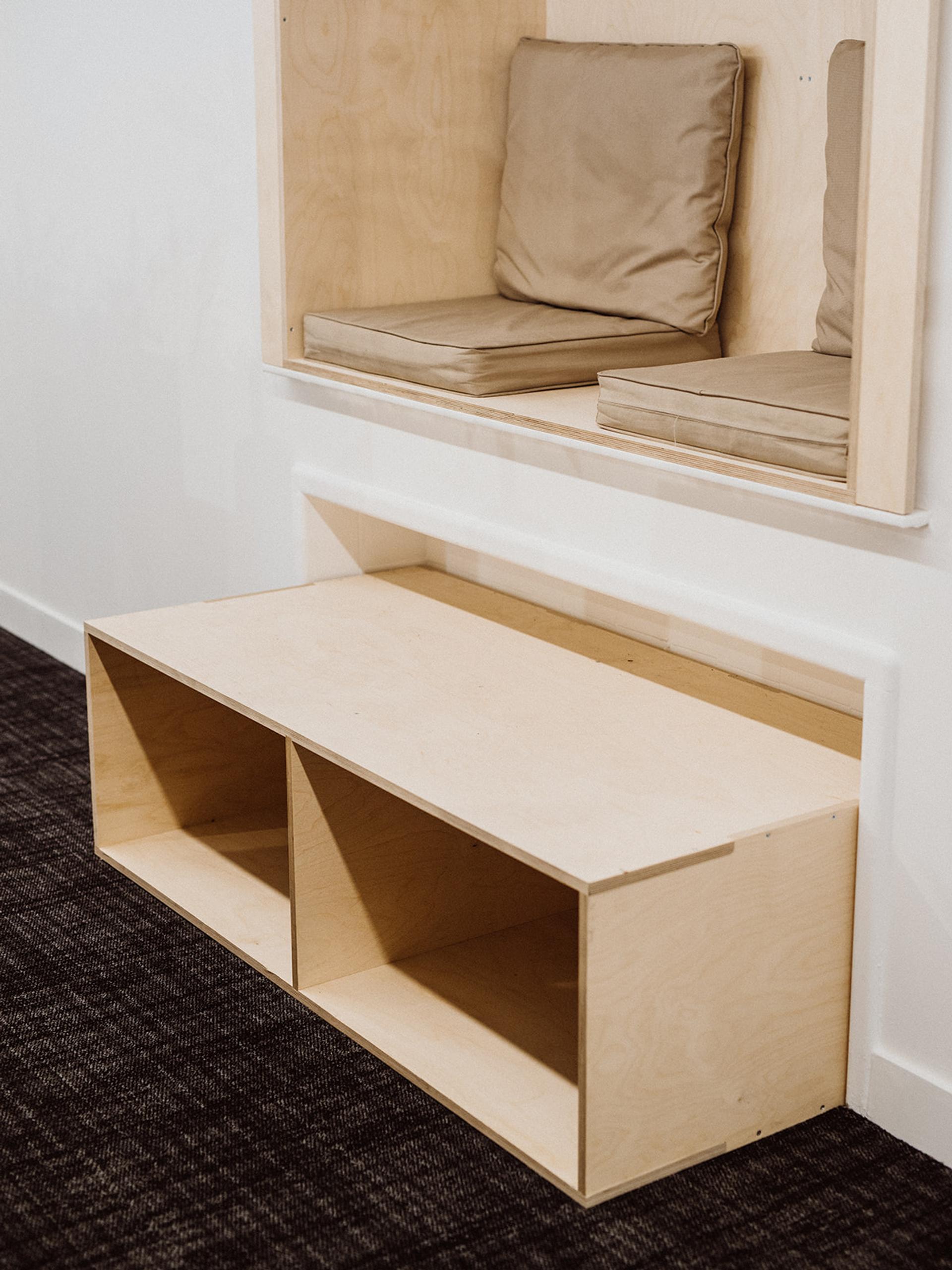 A modern wooden bench with open cubby storage sits against a white wall. Above it, a built-in nook features beige seat cushions and backrests. This modular design highlights sustainable timber construction on a dark brown carpeted floor.