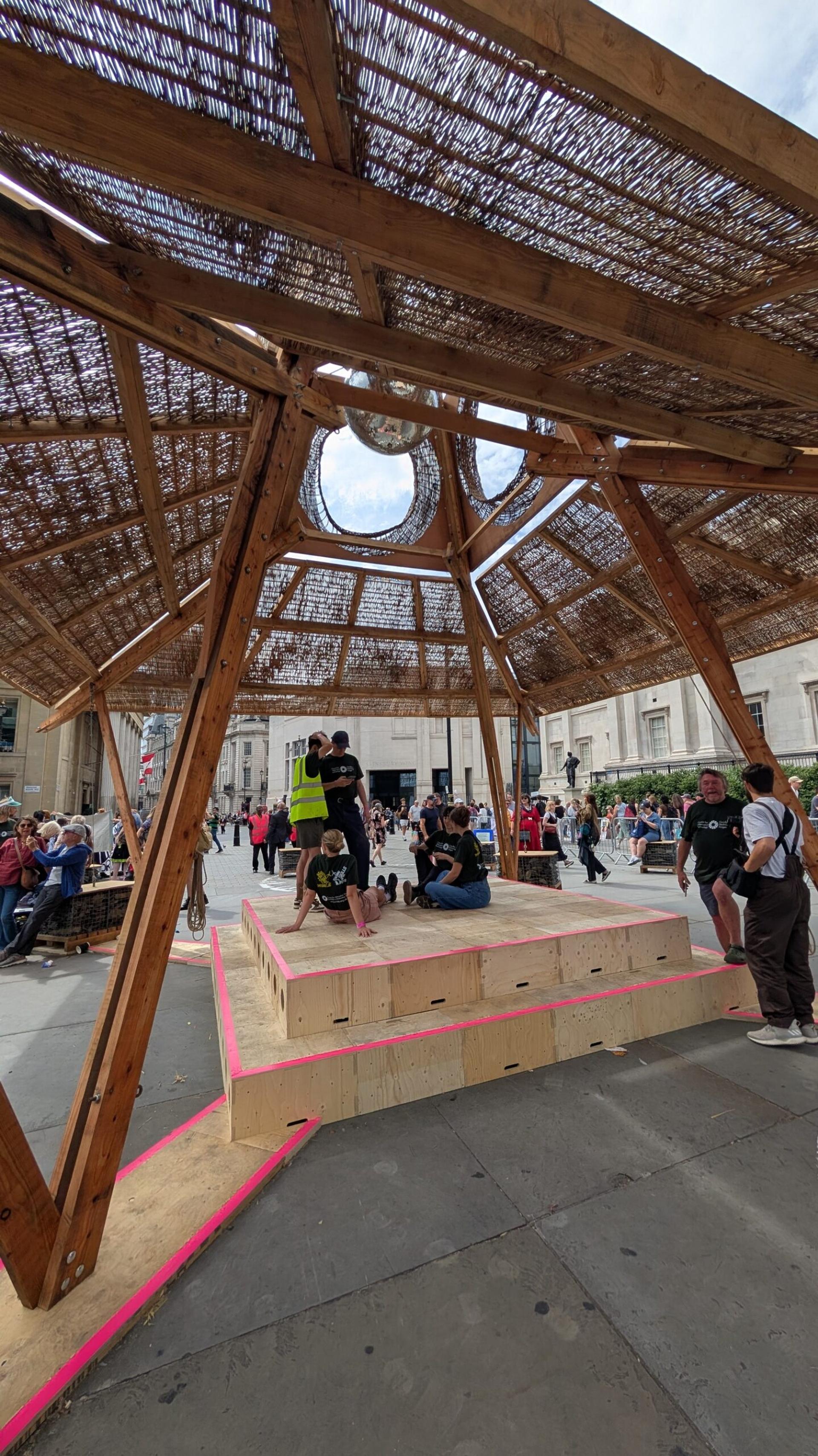 People sit and gather on a wooden platform under a circular wooden shelter with a woven roof, showcasing upcycling and reuse principles, in a busy outdoor public square. Buildings and more people are visible in the background.
