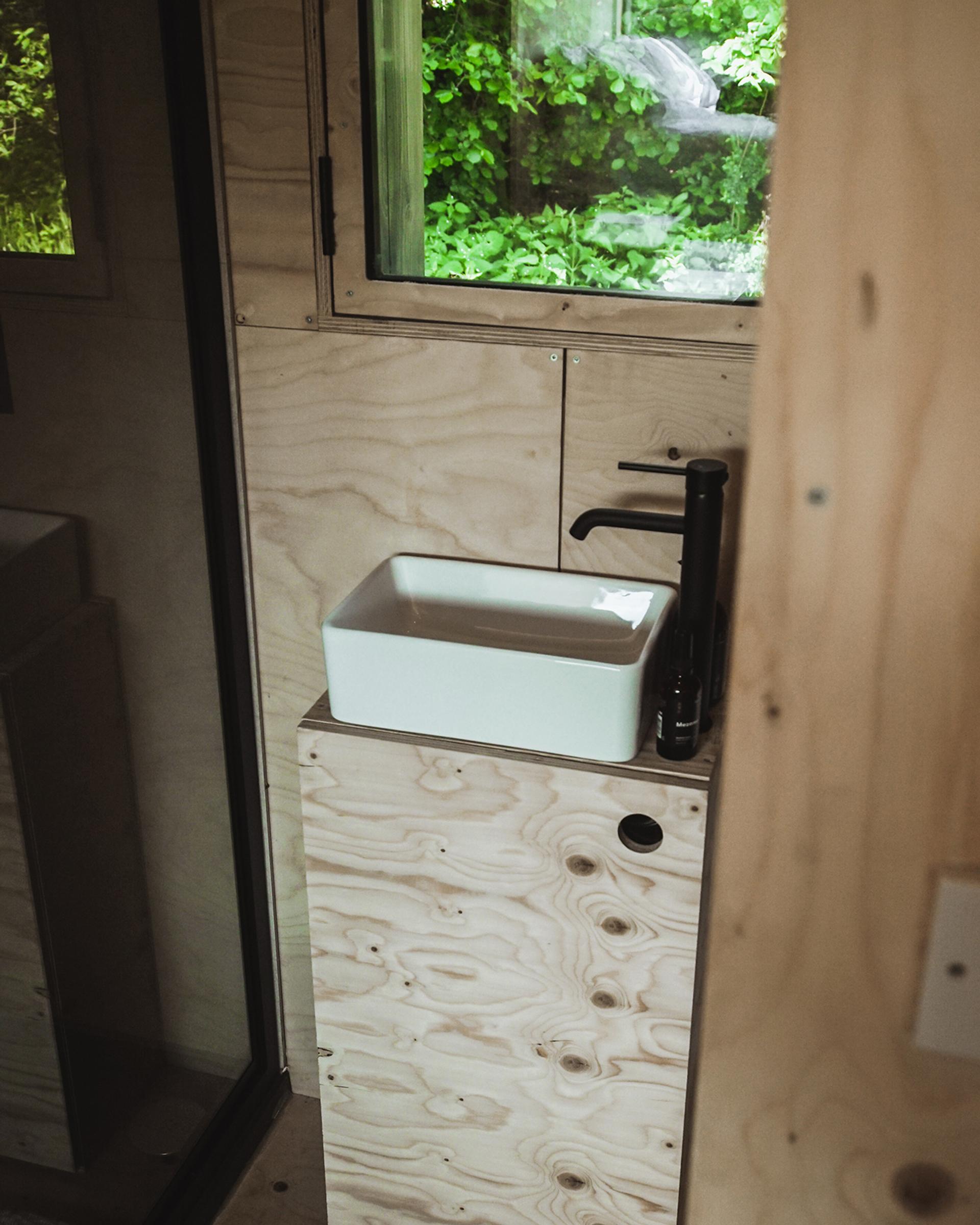 A small modern bathroom with light plywood walls, a rectangular white basin on a wooden vanity, and a black tap—showcasing reuse and circular economy principles—with a window framing greenery outside.
