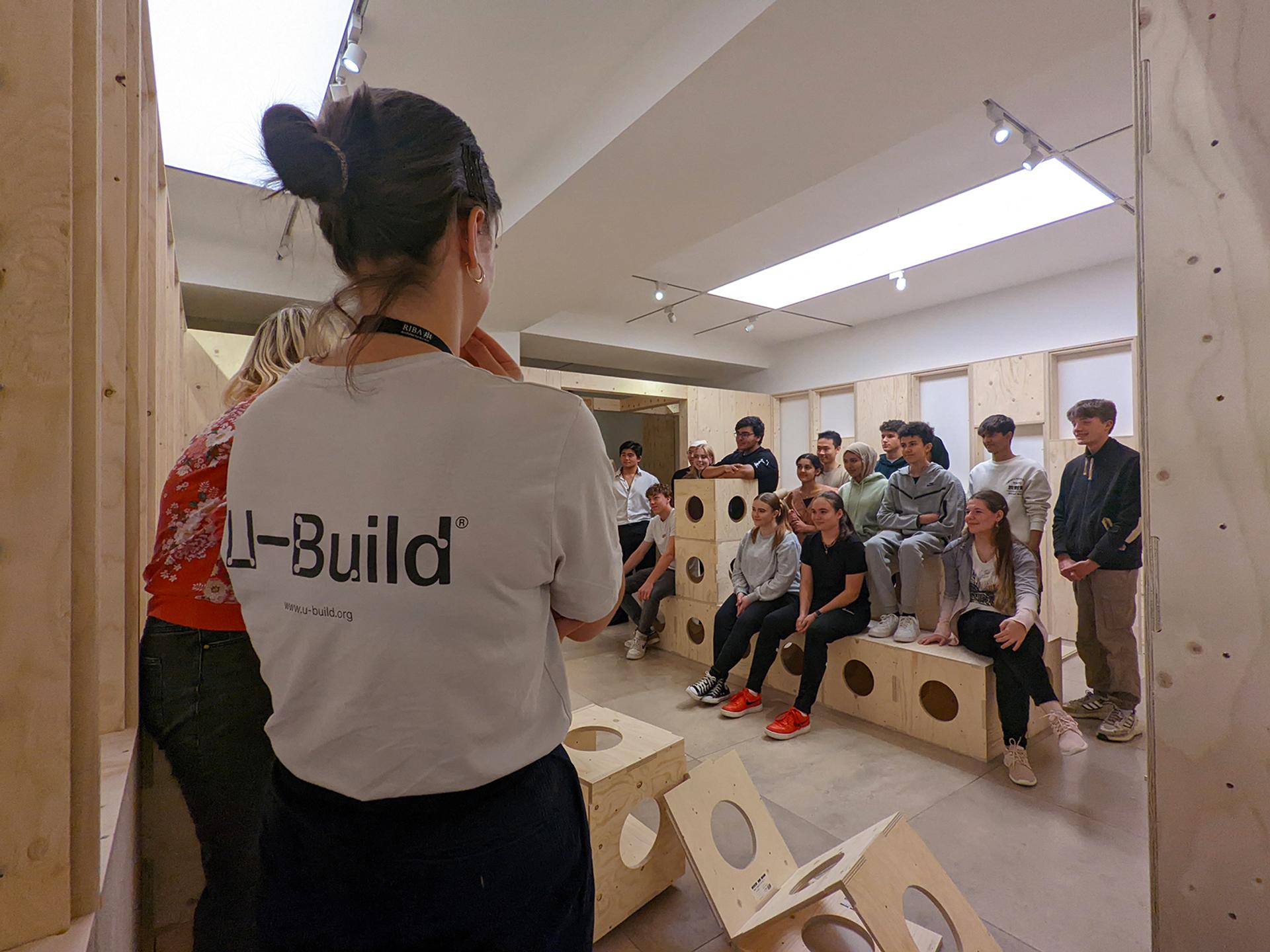 A group of people sits and stands on modular wooden furniture in a modern room while listening to a woman in a white “I-Build” t-shirt speaking at the front. The setting appears to be a workshop or presentation.