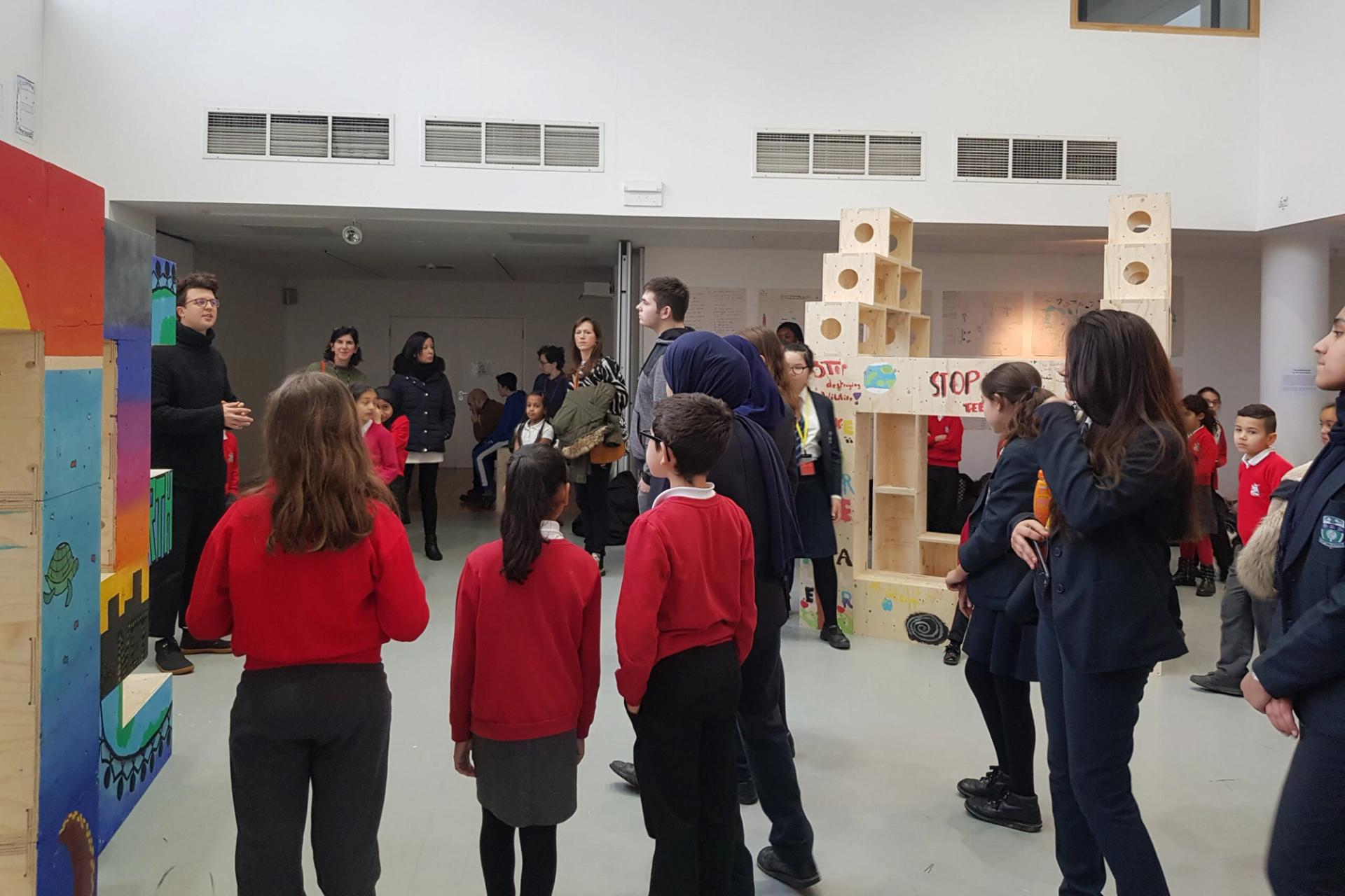 A group of children and adults are gathered in a brightly lit room, some wearing red jumpers and others in dark uniforms, listening to a man speak near colourful wooden displays made from upcycled materials and tall structures.