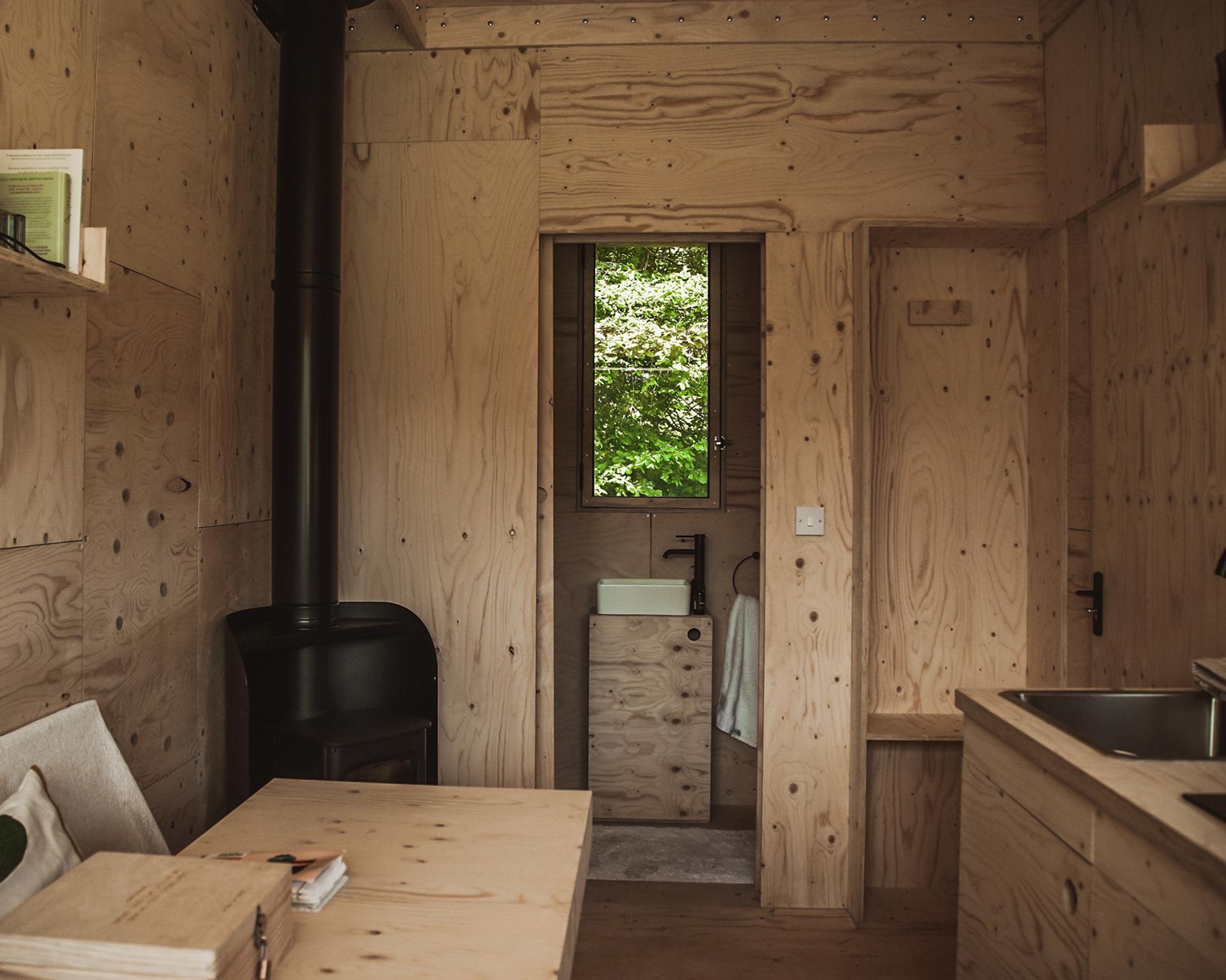 A small, minimalist cabin interior with light plywood walls made from sustainably sourced materials, a wood-burning stove, a simple sink under a window, a worktop, and a wooden table with shelves. Green foliage is visible through the window.