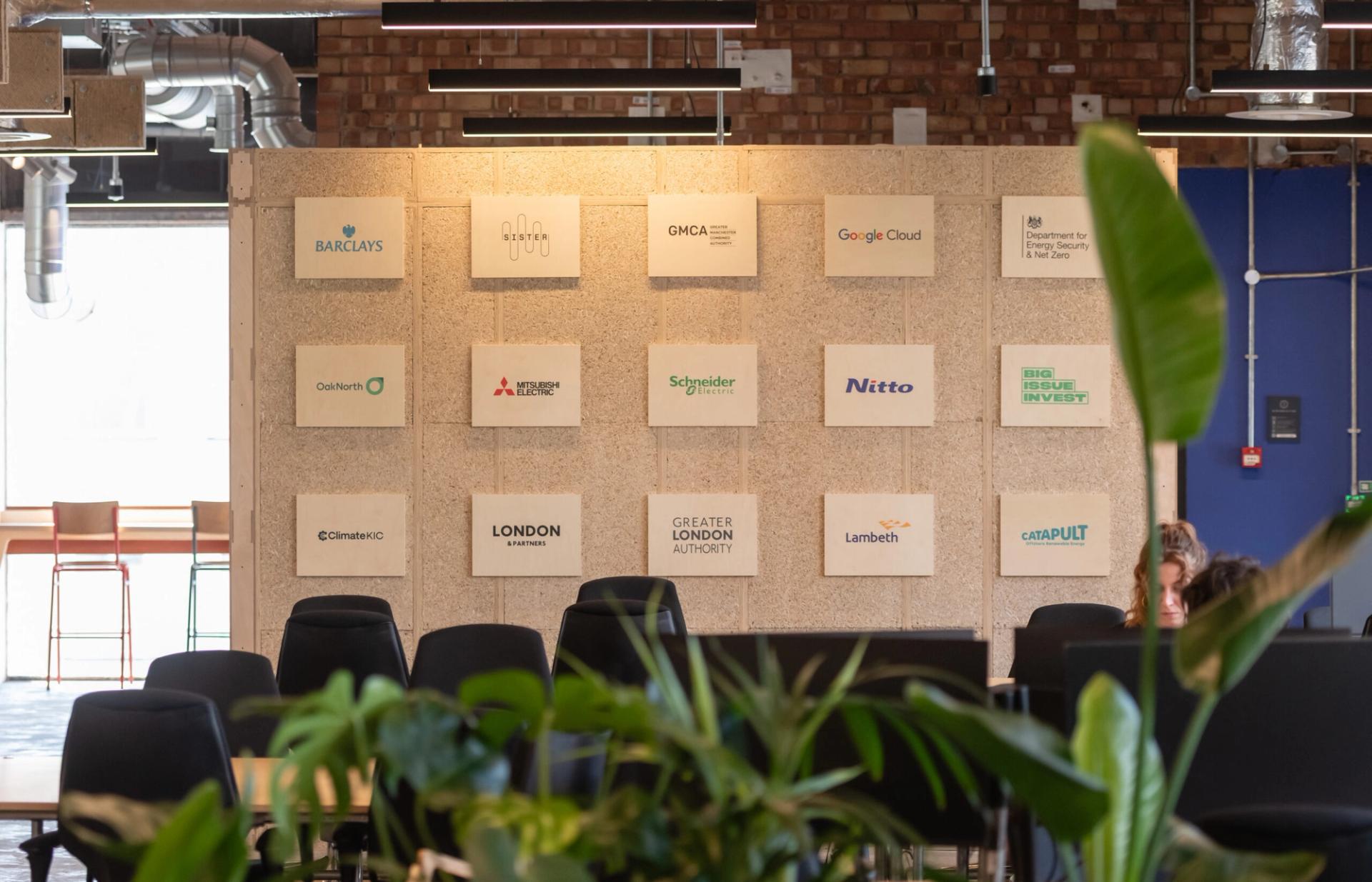 A modern Manchester office space with a wall displaying various company logos, including Barclays, Google Cloud, and Sustainable Ventures. Green plants and empty chairs are visible in the foreground.