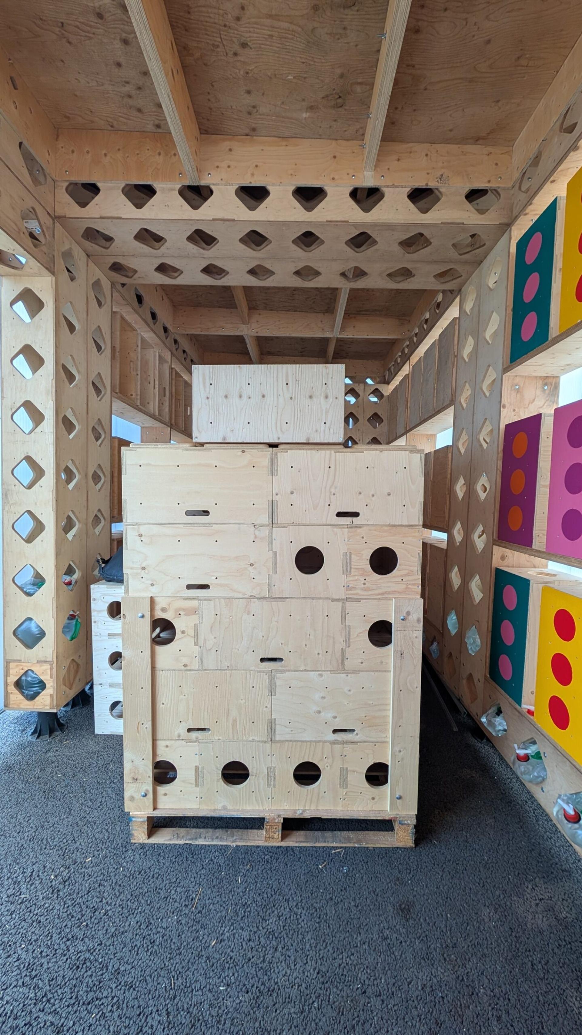 Stacks of U-Build wooden boxes with circular holes are arranged inside a wooden structure featuring cut-out diamond shapes on the walls. Colourful panels with large dots are visible on the right side of the image.