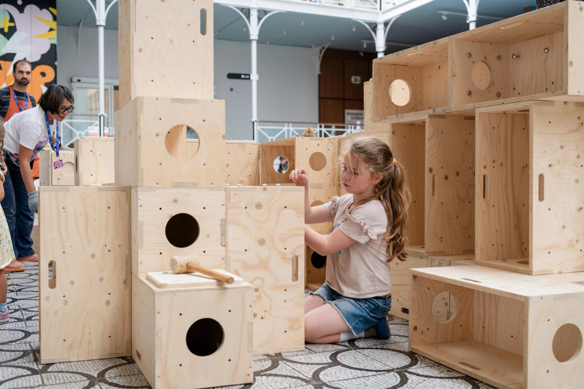 A young girl kneels on the floor, building with large, light U-Build wooden blocks that have circular holes. Other people are visible in the background inside a spacious, bright room.