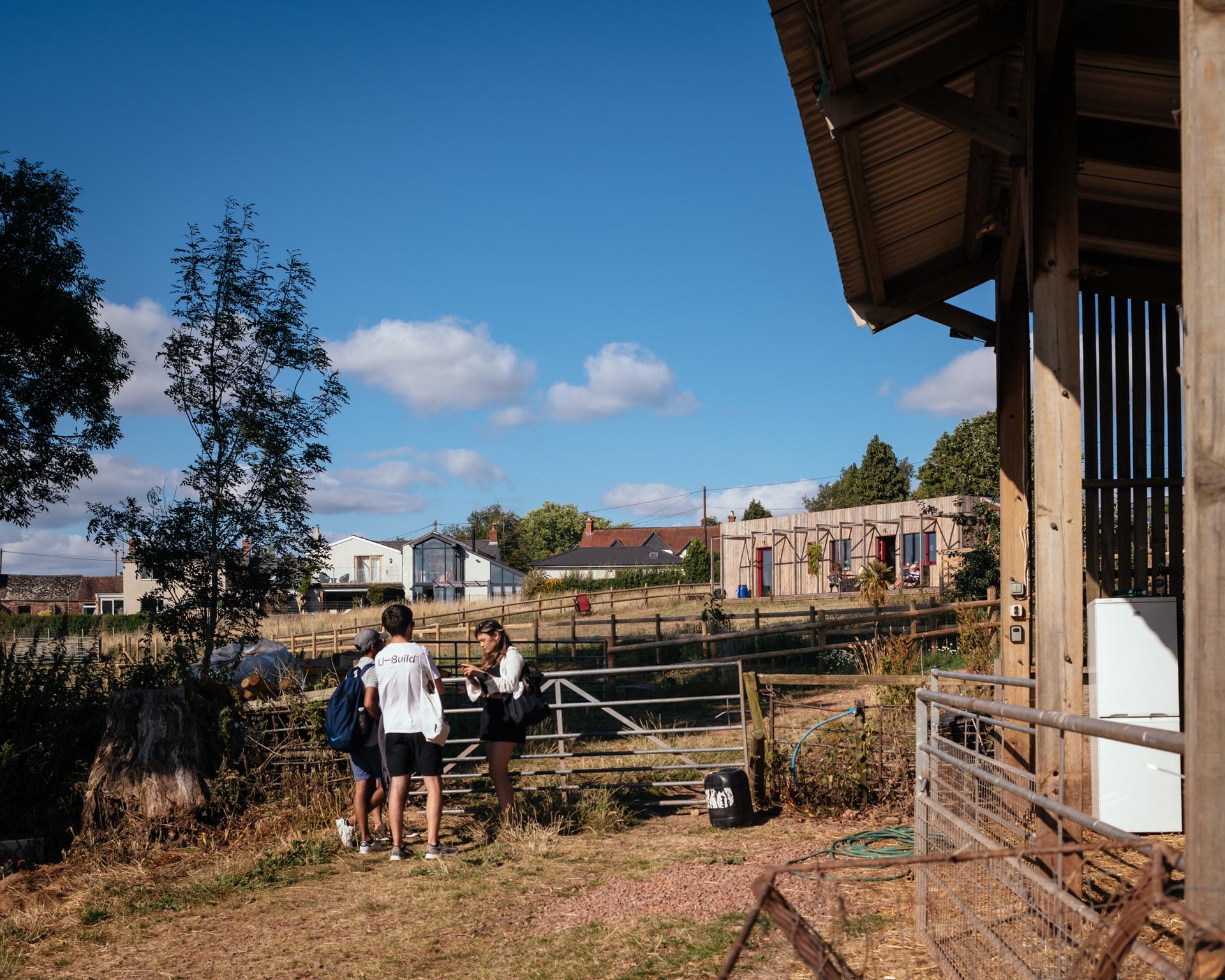 Three people stand and talk near a metal gate on a sunny farm. With houses and barns featuring sustainable timber construction in the background, a clear blue sky and scattered clouds stretch above, whilst part of a wooden shed is visible on the right.