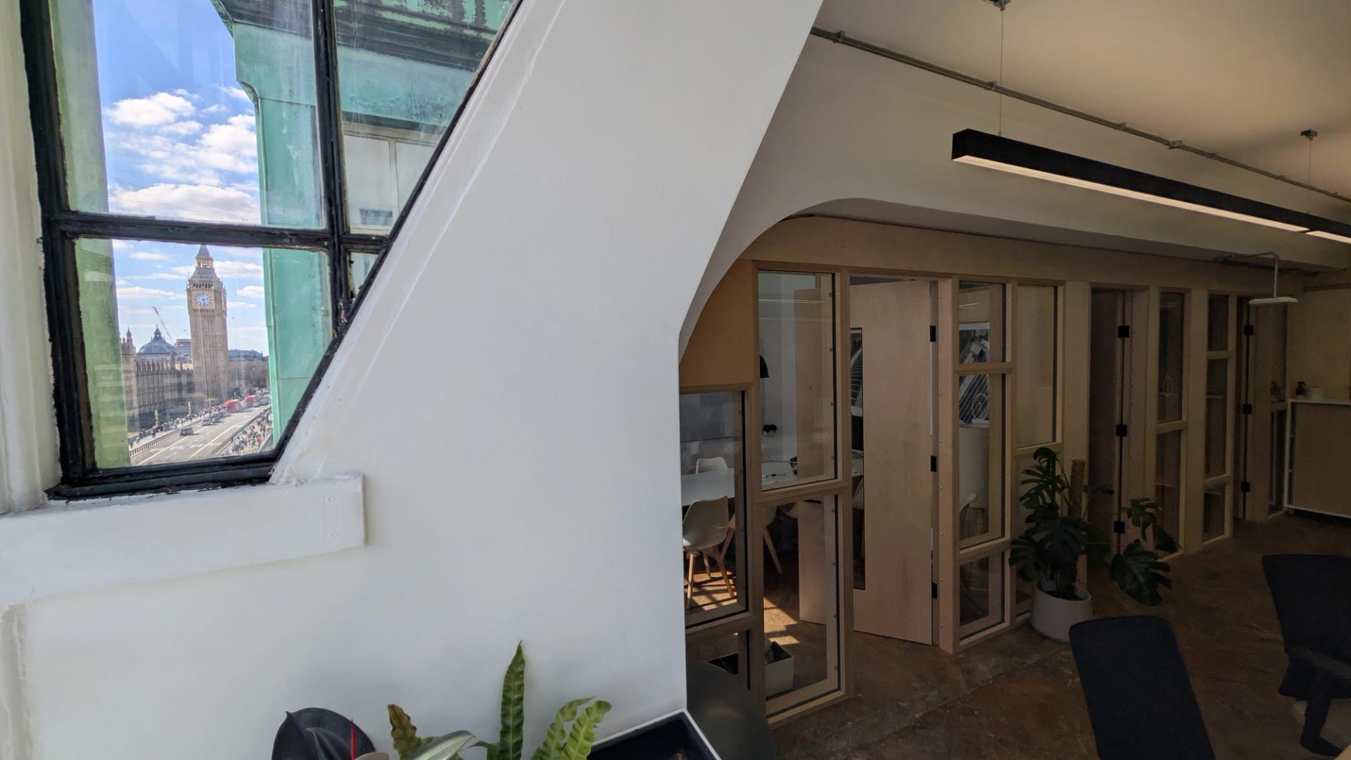 A bright office with glass meeting rooms, potted plants, and birch plywood accents, plus a large arched window showing Big Ben and the Houses of Parliament under a blue sky.