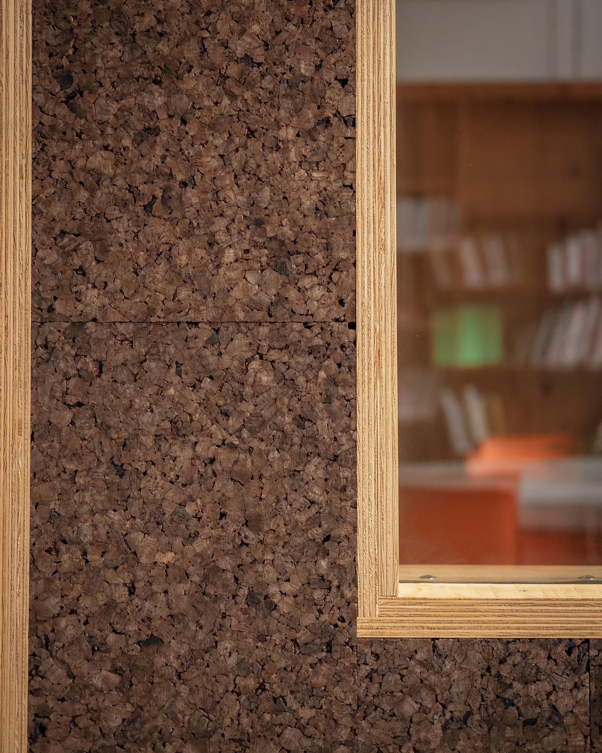 Close-up of a sustainable cork wall with a light spruce-framed window on the right, revealing blurred shelves and books in the background.