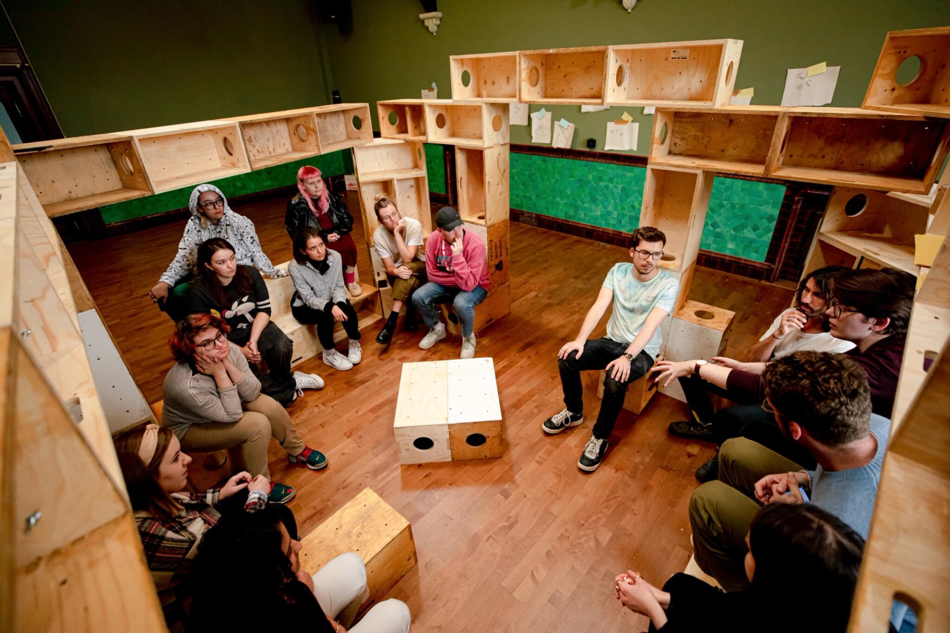 A group of people sit in a circle on spruce stools and boxes inside a room with green walls and wooden flooring, engaged in conversation. Timber construction shelves with papers attached line the sustainable space.