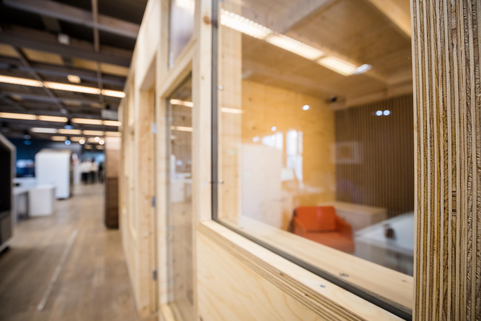 A close-up view of a modular wooden and glass office partition, with a glimpse of a modern, open office space in the background featuring plywood walls, bright lighting, and contemporary furniture.