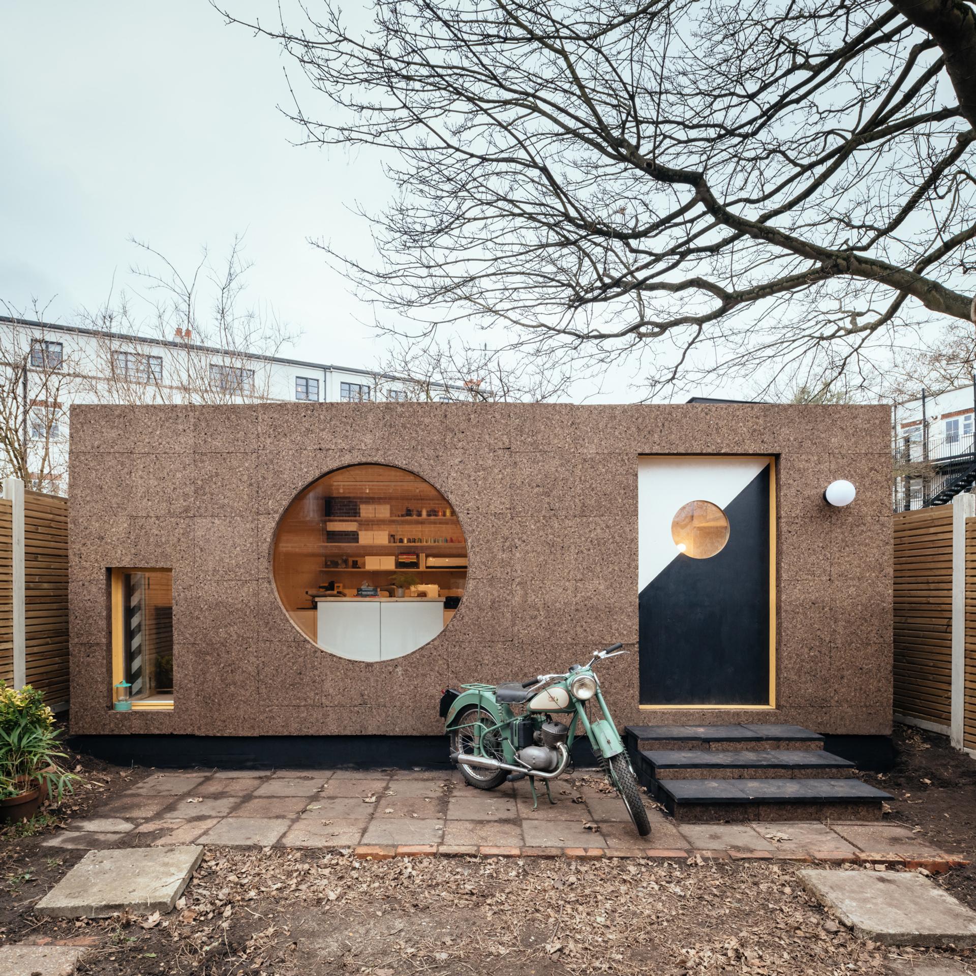 A modern, box-shaped tiny house with a cork exterior promotes sustainability, featuring large circular and rectangular windows and a geometric door. An old green motorbike parked in front highlights creative reuse; leafless tree branches frame the scene.