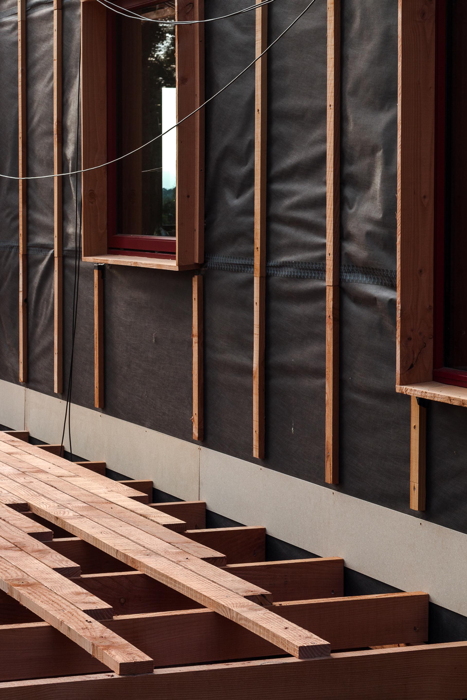 Exterior wall of a building under construction with exposed timber framing, black weatherproof barrier, visible window frames, and wooden beams laid out for a deck or floor structure, highlighting sustainability in modern building practices.