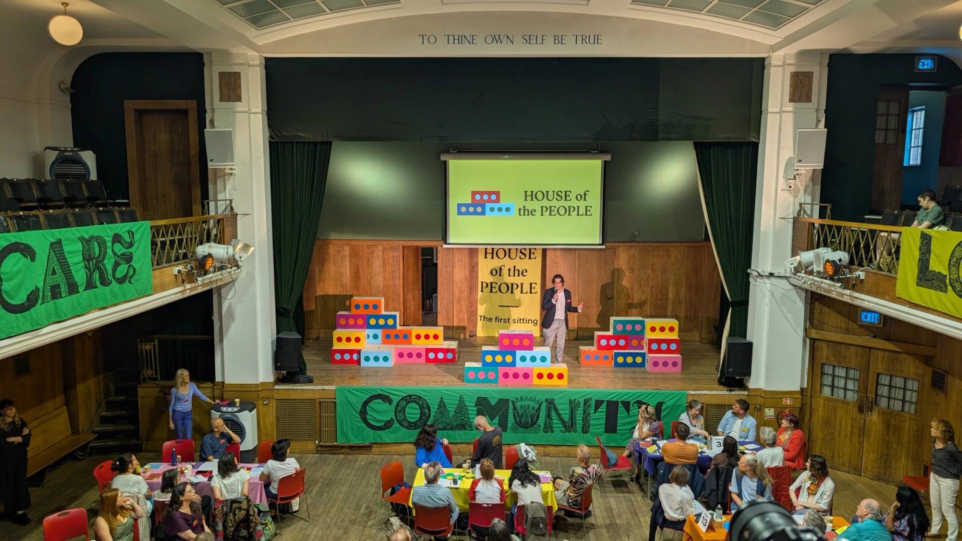 A community hall event with people seated at tables. A person speaks on stage beneath a “HOUSE of the PEOPLE” sign. Bright banners reading “CARE,” “COMMUNITY,” and “Sustainability” hang from the balcony.