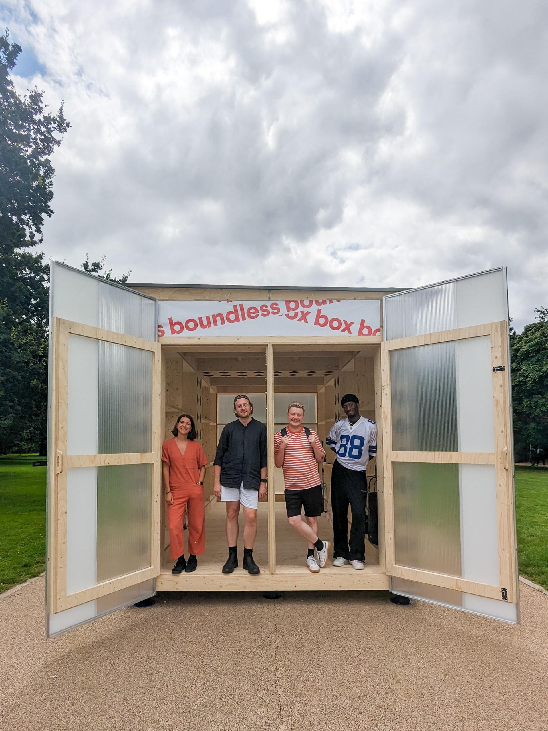 Four people stand smiling inside a modular timber construction with open doors on a park path. The sign above reads boundless box. Trees and a cloudy sky are visible in the background.