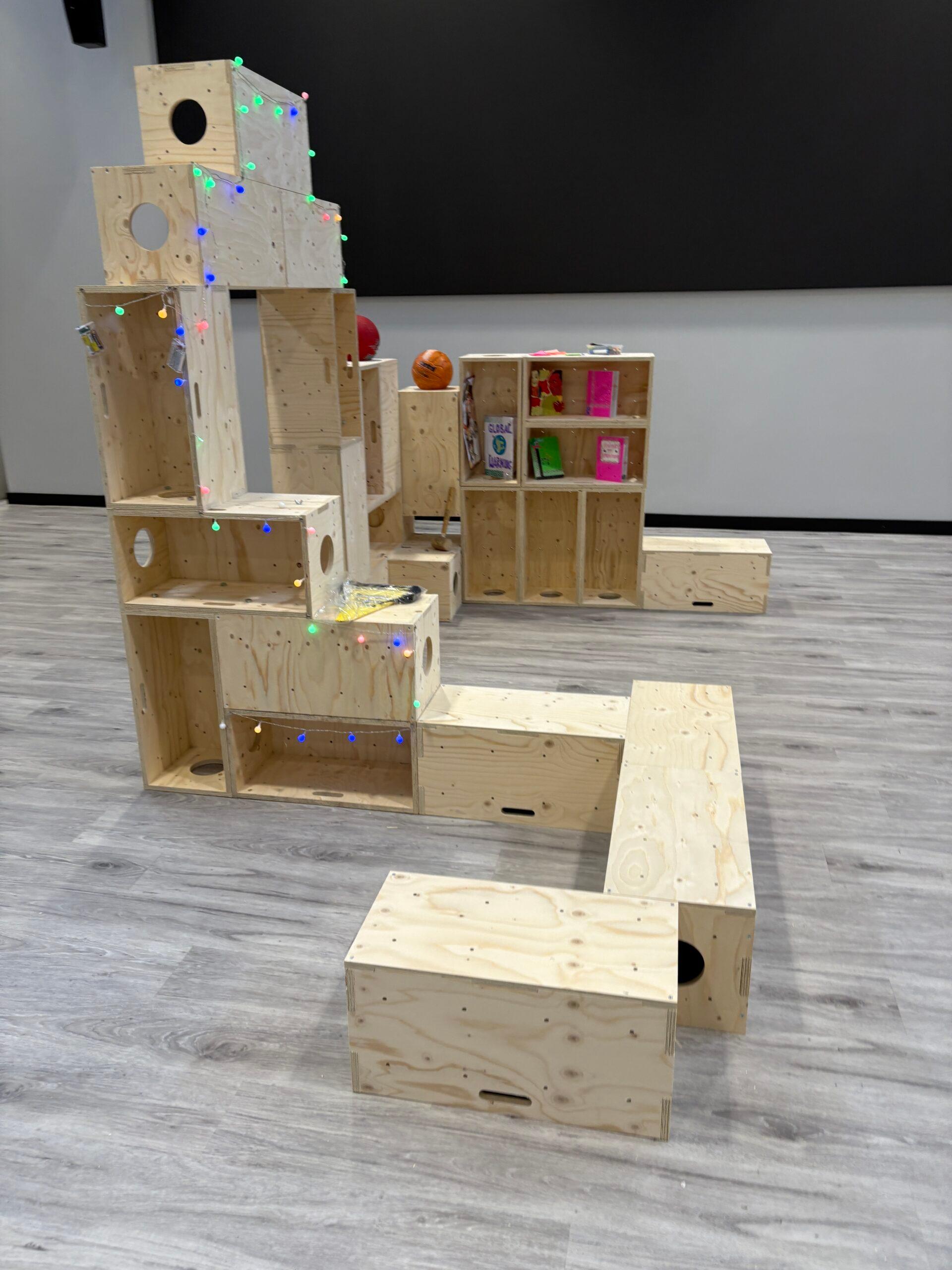 A modular wooden play structure crafted from birch plywood features stacked boxes, some adorned with fairy lights. Shelves hold books and toys, all set on a light wood floor in a modern room.