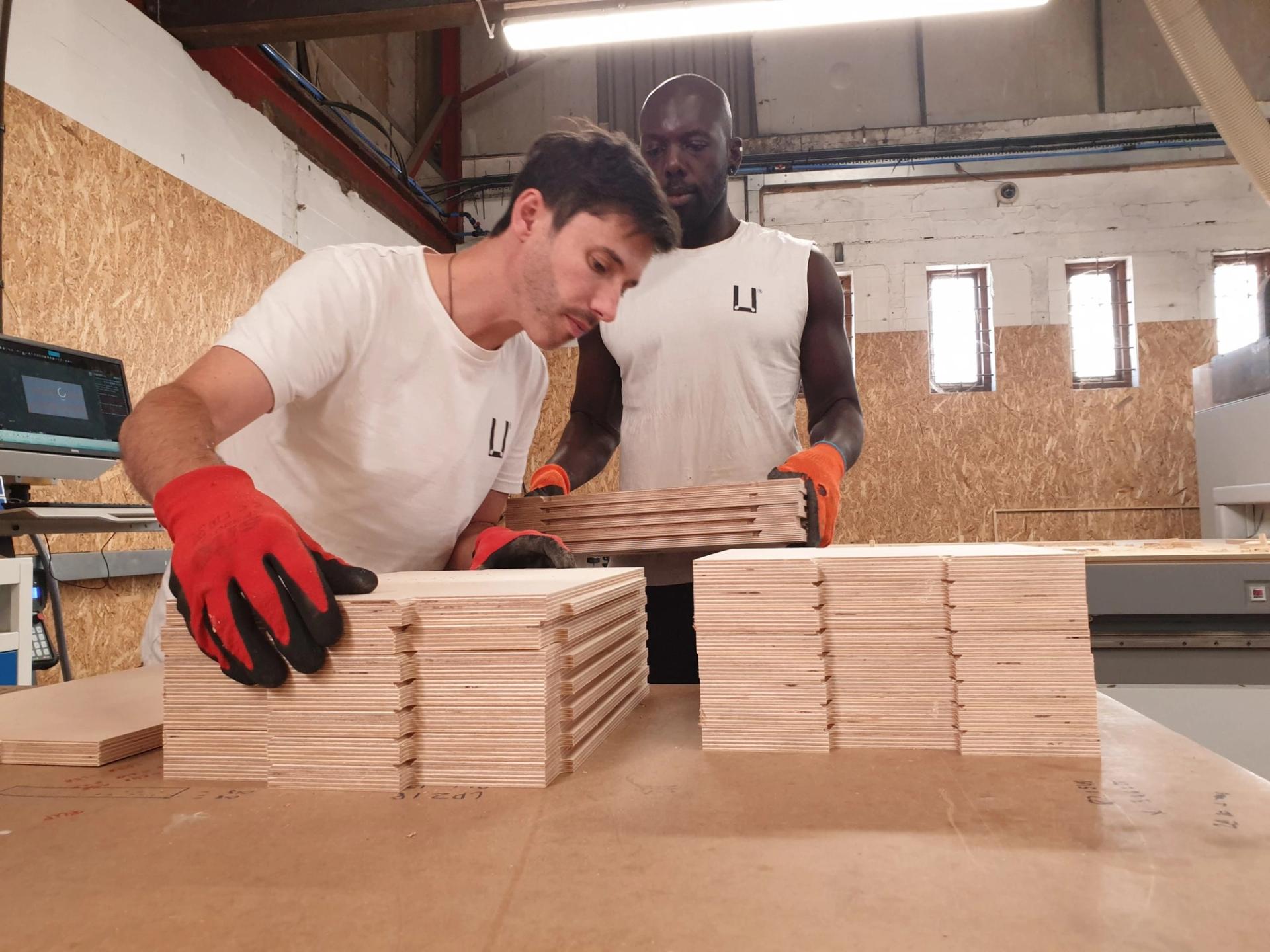 Two men wearing white shirts and orange gloves stack rectangular birch boards on a table in a workshop with a wood-panelled wall and computer equipment in the background.
