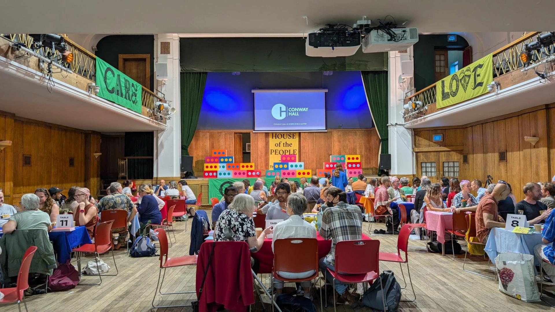 A large room with groups of people seated at tables, engaged in discussions. Colourful banners and blocks decorate the stage, promoting the circular economy, while a screen displays Conway Hall. The atmosphere is lively and collaborative.