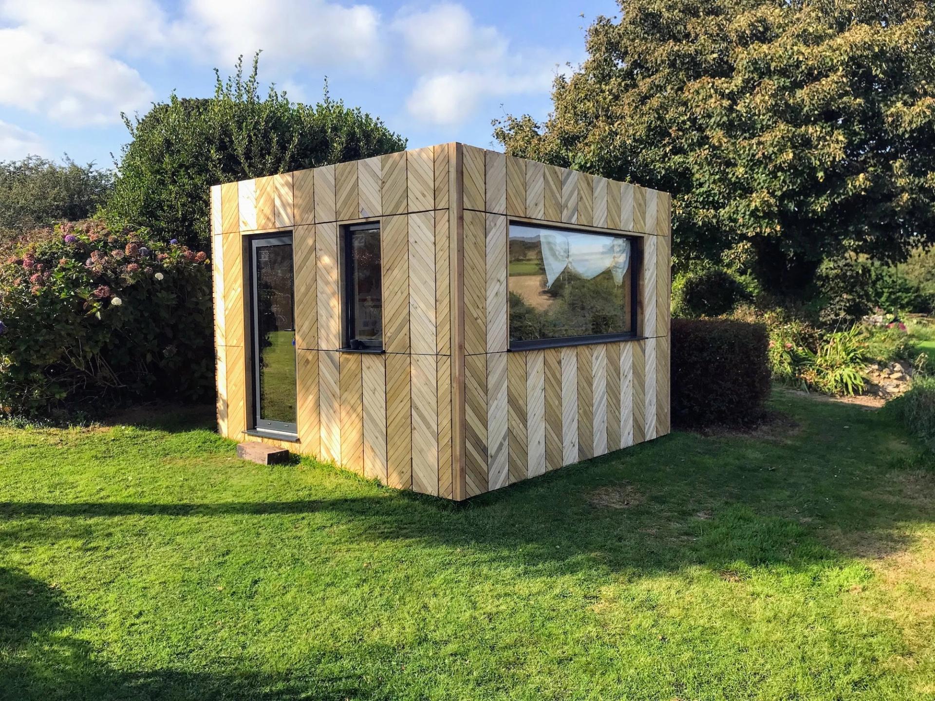 A small, modern spruce garden shed with large windows stands on a grassy lawn, surrounded by green bushes, trees, and flowering plants on a sunny day. Its sustainable timber construction blends beautifully with the natural setting.