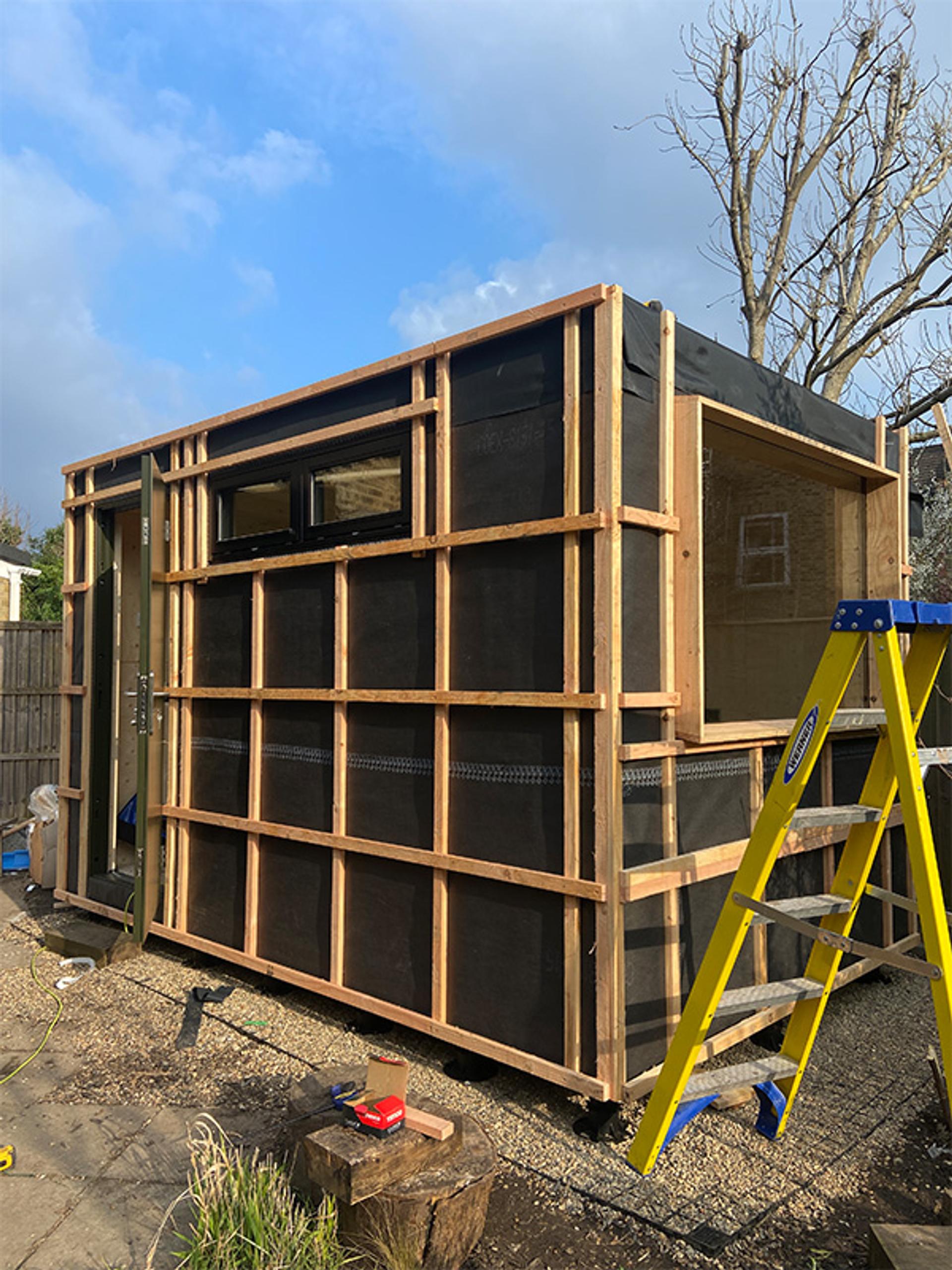 A small modular wooden structure under construction outdoors features exposed spruce framing and black weatherproof sheeting. A yellow ladder stands nearby, with building materials scattered on the ground. Trees and fencing are in the background.