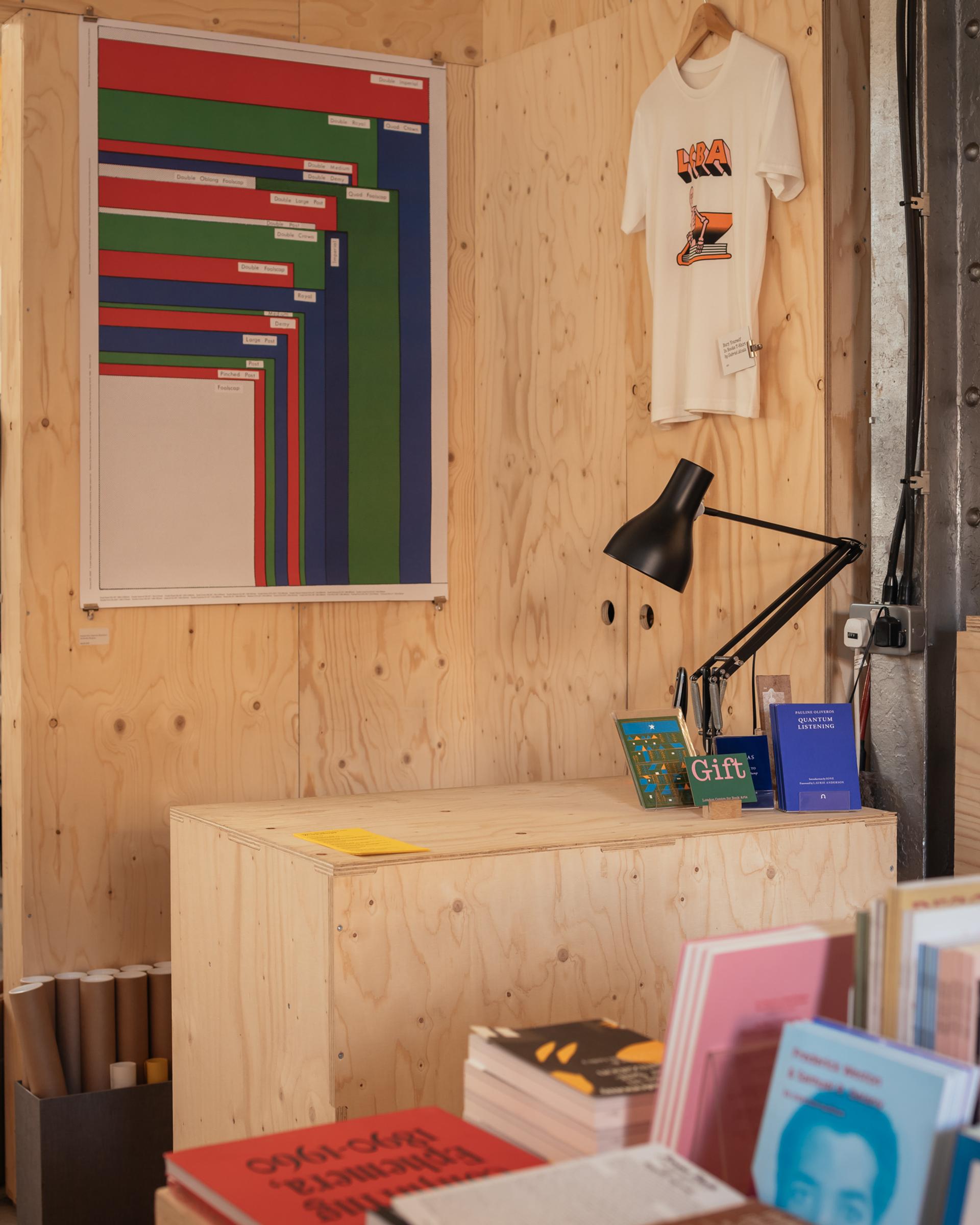 A small spruce-wooden shop counter with books, a desk lamp, and a wall-mounted colourful chart. A white graphic T-shirt hangs above the counter, with more books and posters displayed in this cosy modular space.
