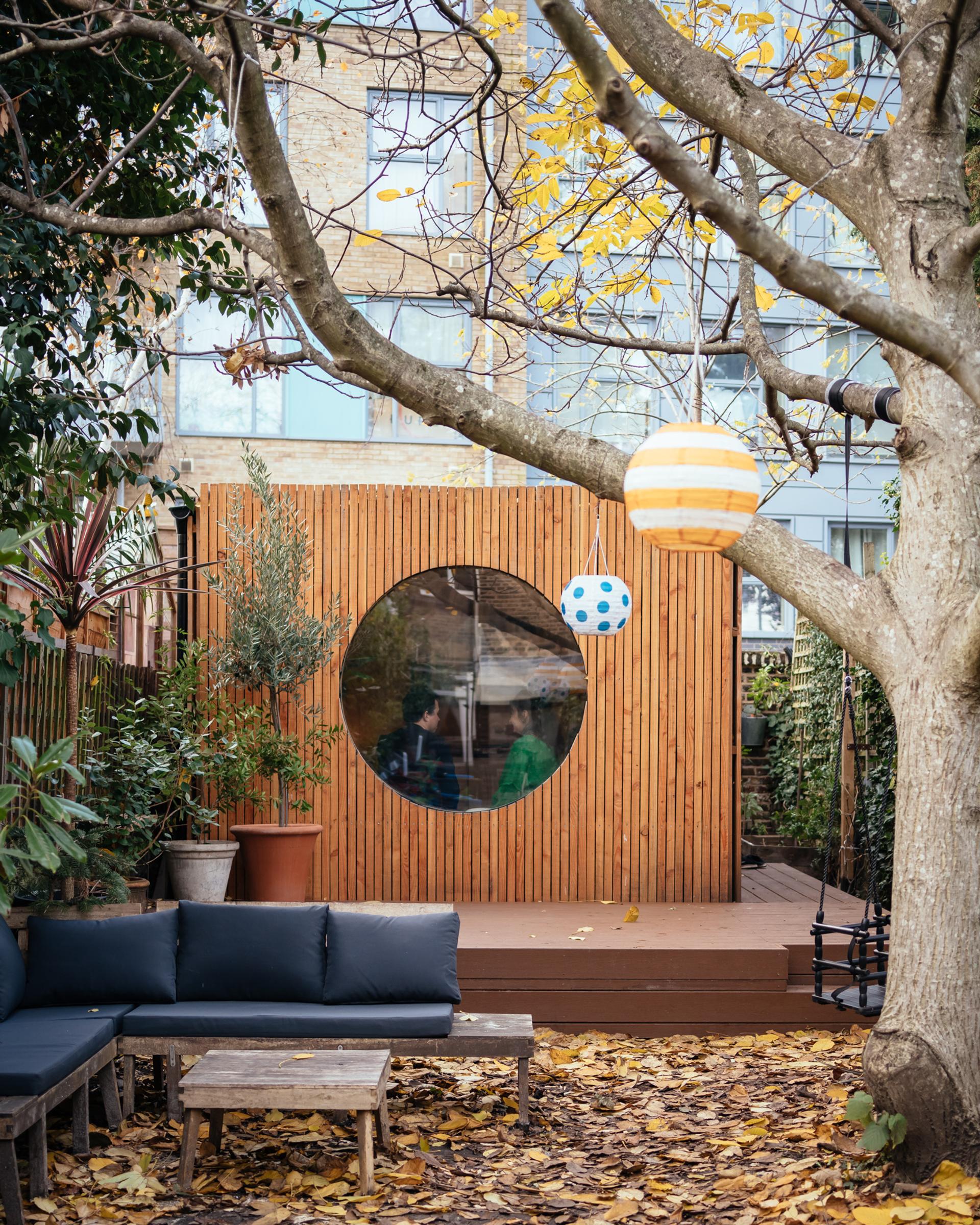 A cosy back garden with a spruce wood decking, circular window, potted plants, blue cushioned seating, a coffee table, and autumn leaves scattered on the ground. Two round lanterns hang sustainably from a tree branch in the foreground.