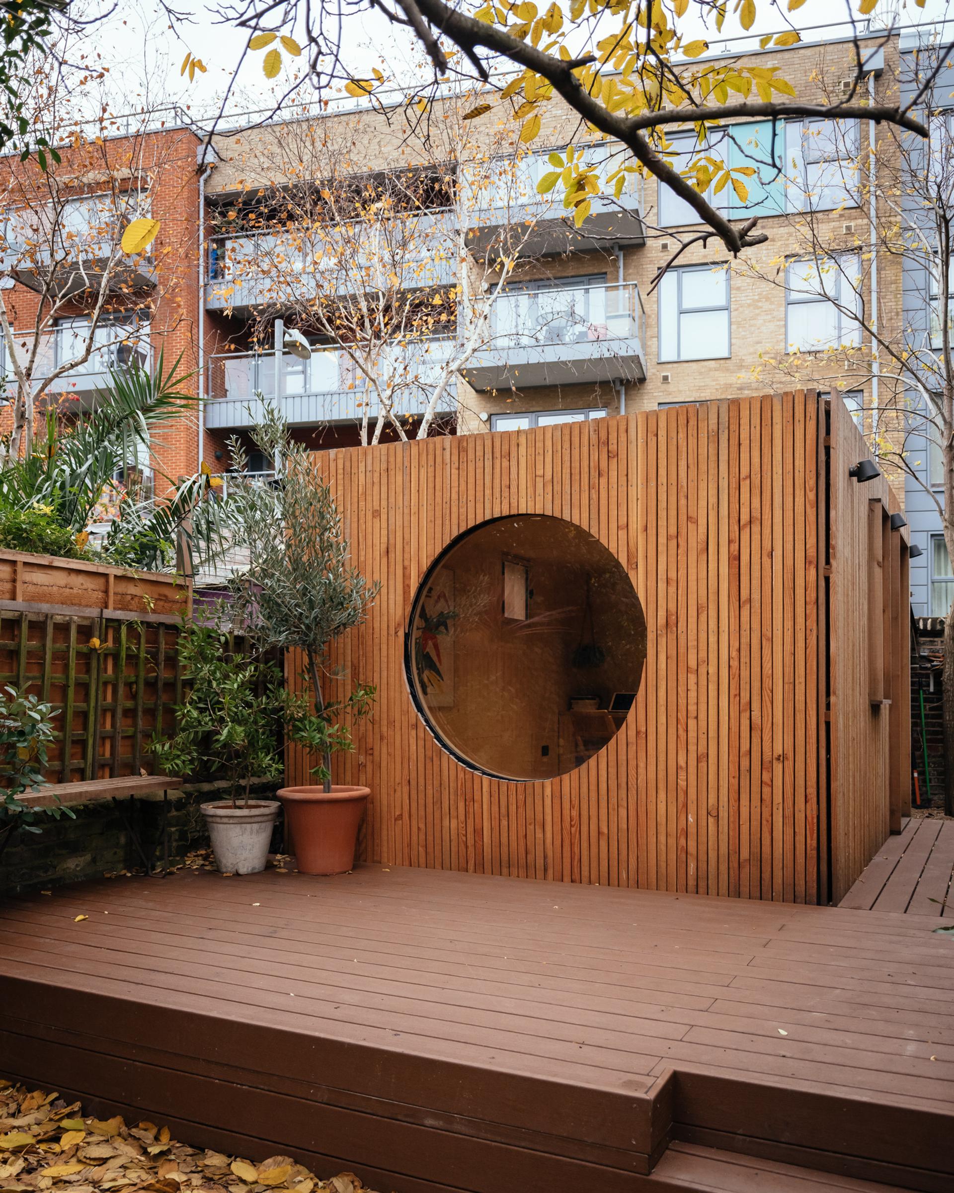 A modern birch plywood garden studio with a large circular window sits on a raised wooden deck in a back garden, surrounded by potted plants and autumn leaves, with blocks of flats in the background.
