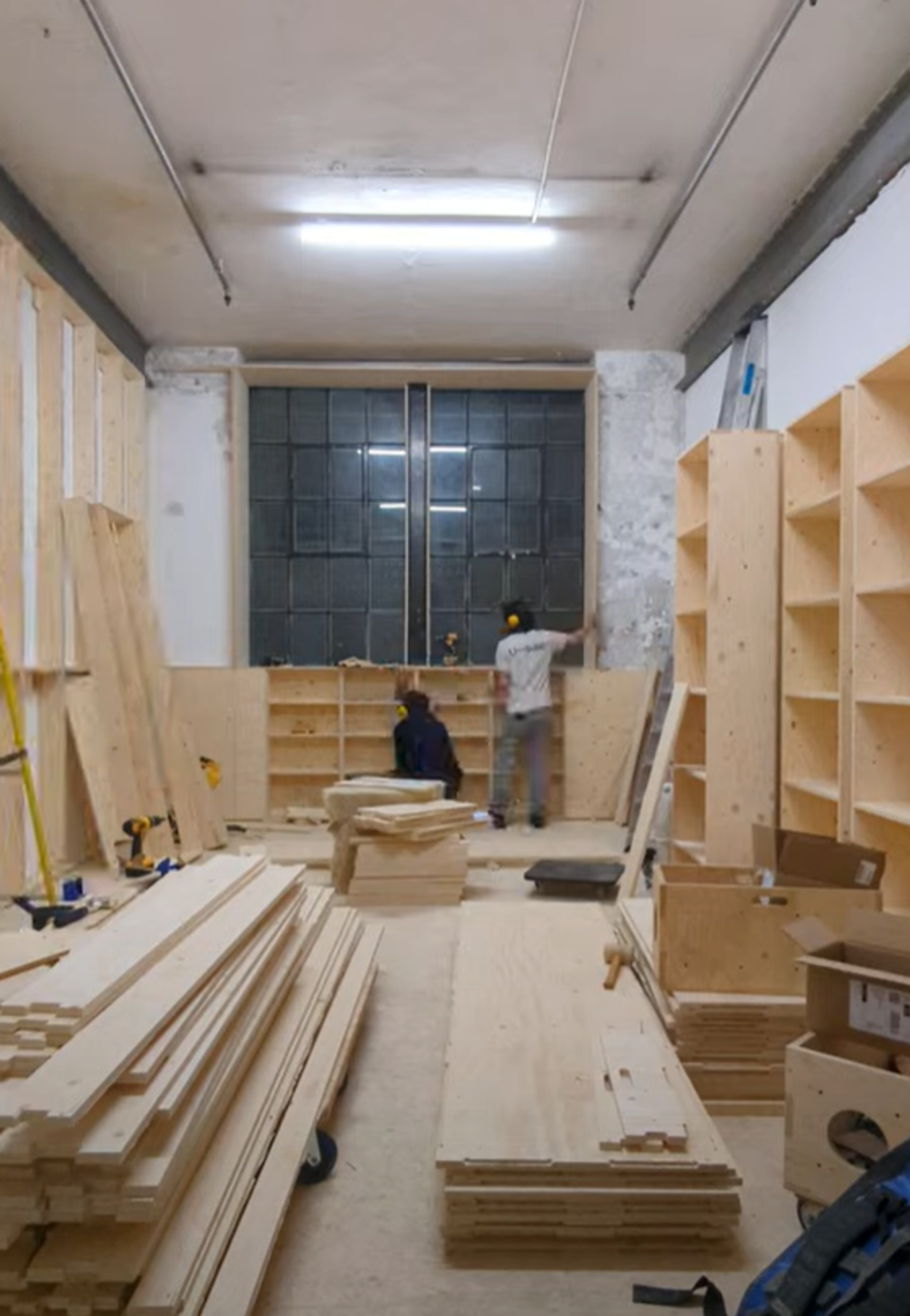 Two people work in a workshop assembling modular wooden shelves and storing spruce planks. The room is filled with stacks of timber and finished shelving along the walls, with a large window at the back letting in light.