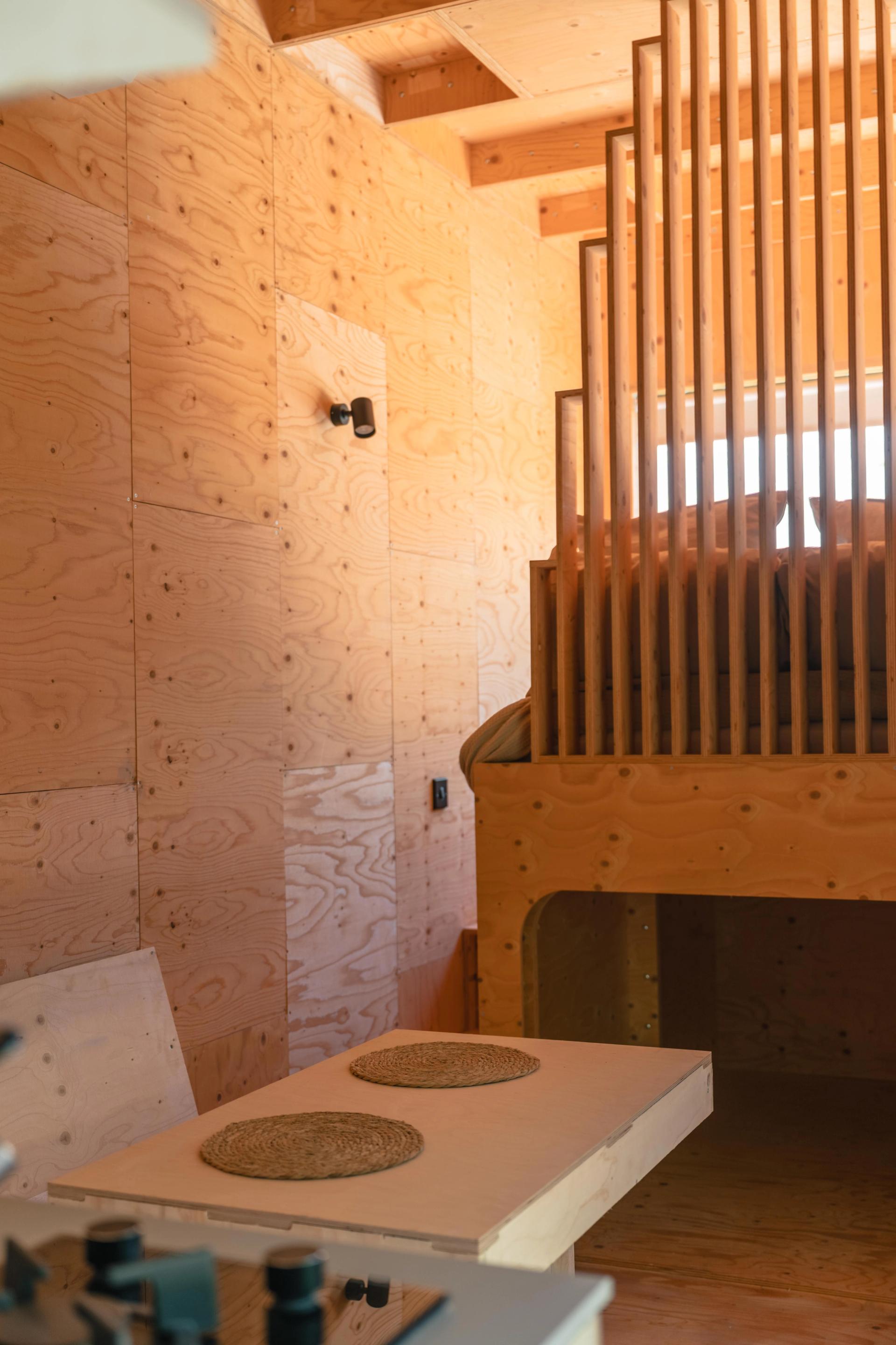 A cosy, minimalist wooden interior with cabin furniture, featuring a lofted bed, vertical slats for railing, a small fold-out table set with two place mats, and natural light streaming in.