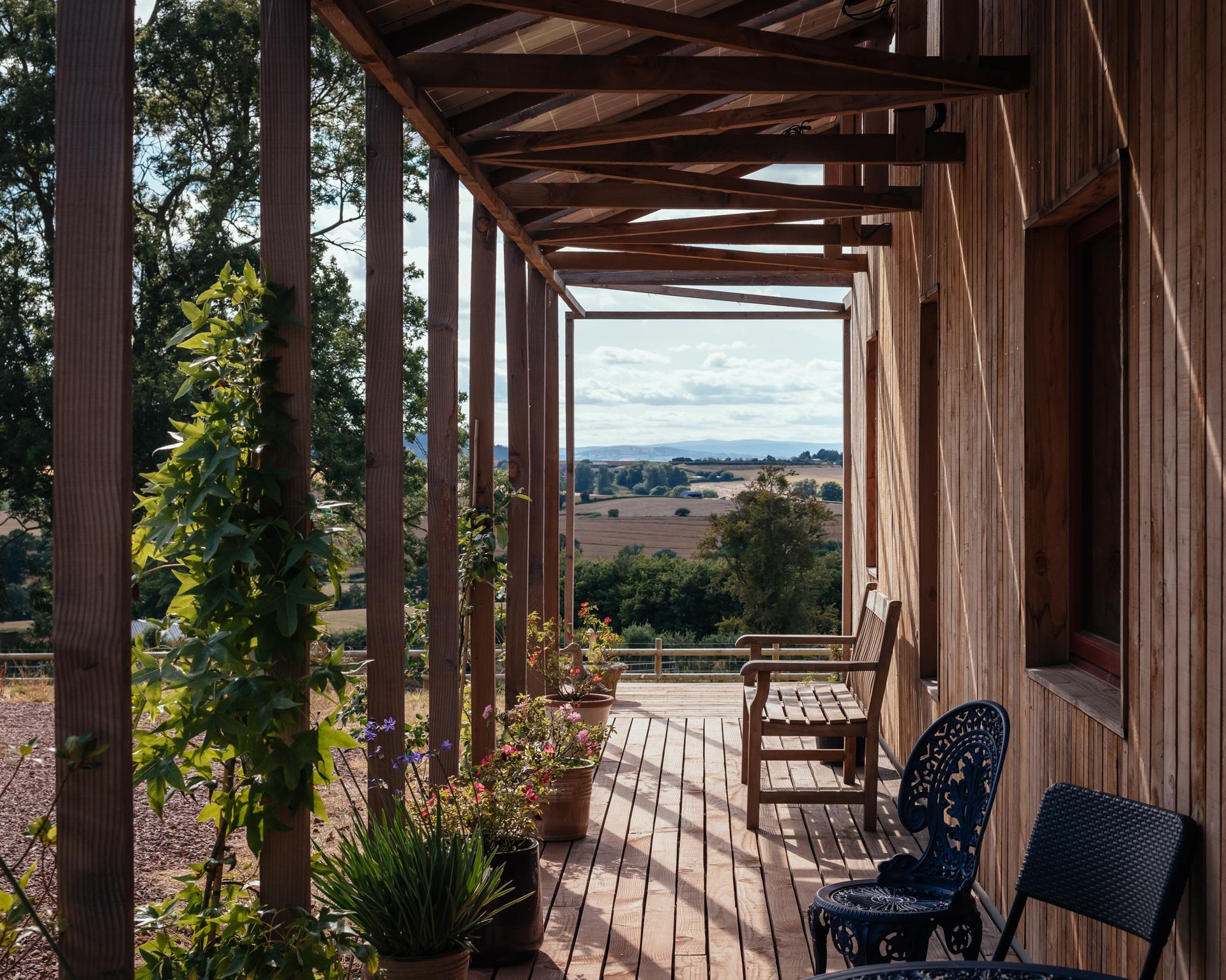 A wooden veranda built with sustainable timber construction features potted plants, a bench, and metal chairs overlooking fields and distant hills under a partly cloudy sky. Sunlight casts gentle shadows across the veranda.