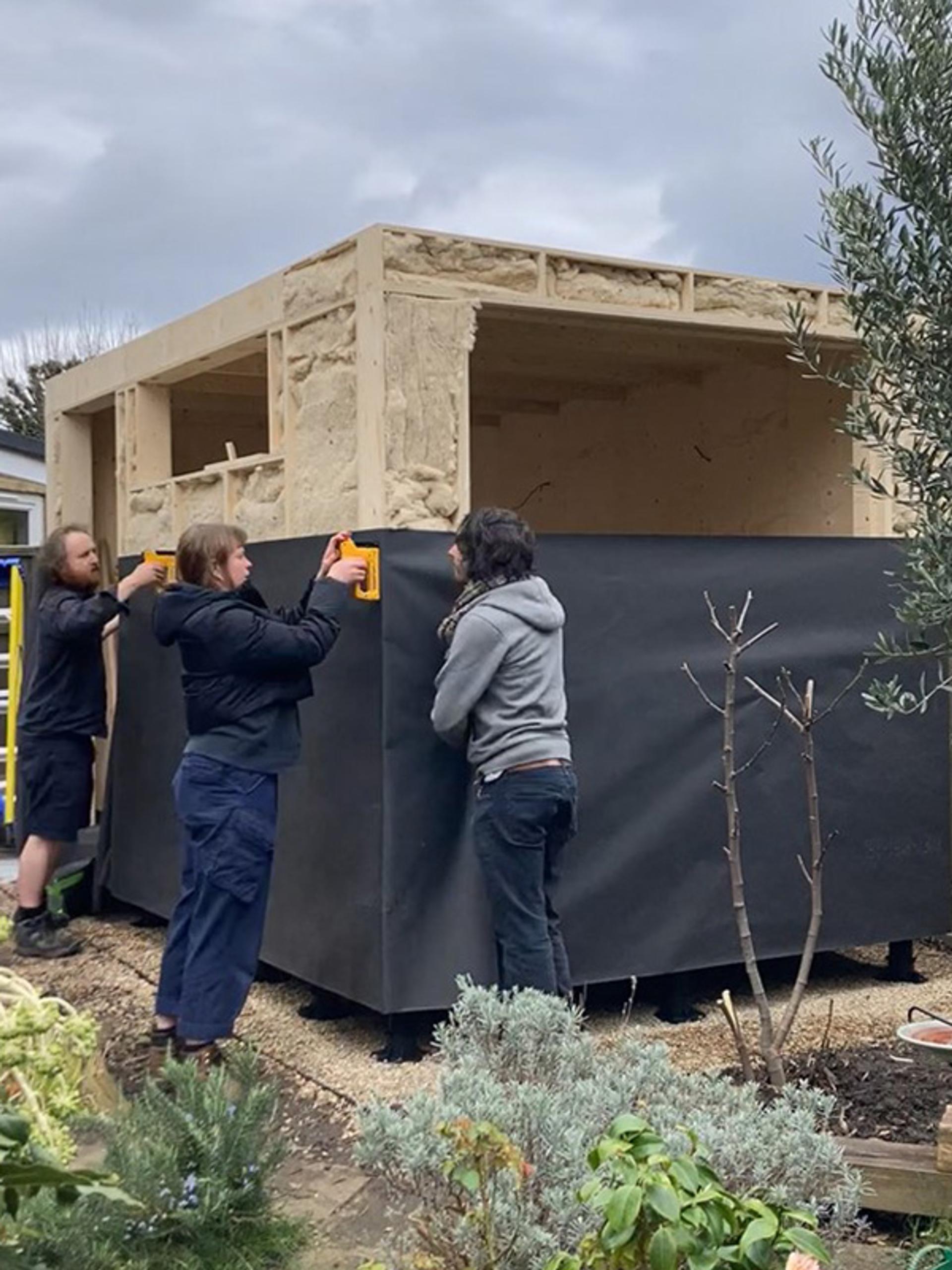 Three people are building a small timber construction, attaching black material to its exterior walls. The structure stands in a garden with various plants, under a cloudy sky.