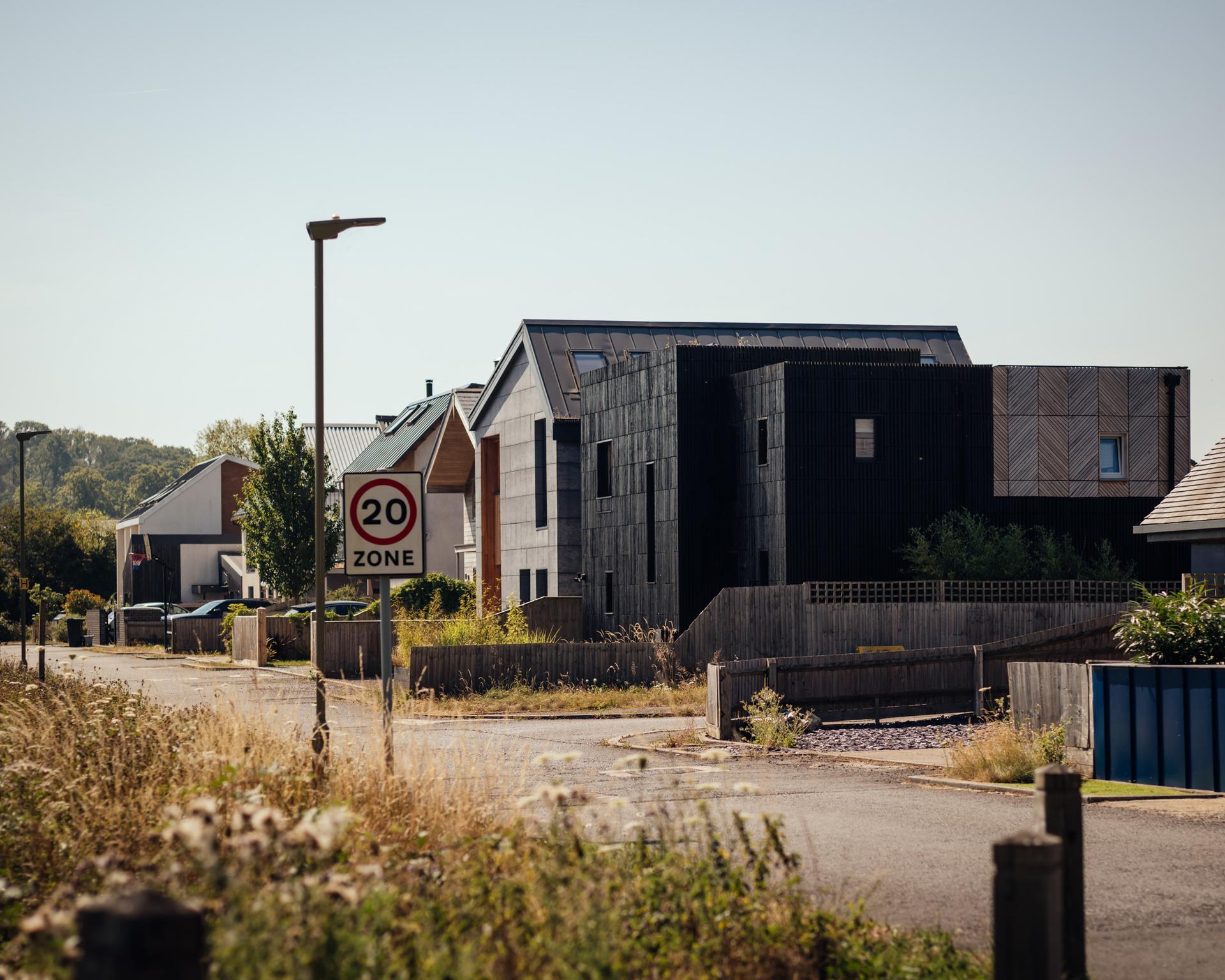 A 20 mph zone sign stands on a suburban street lined with modern homes showcasing box house extension architectural design and black and grey façades.