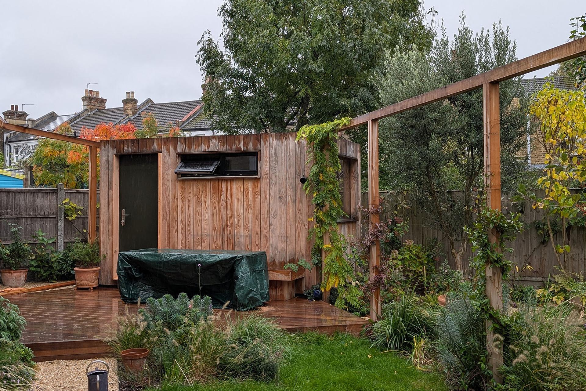 A U-Build wooden garden shed with a dark door and small window sits on a wet wooden decking. Plants climb up trellises and the shed, with a covered object in front. Trees and shrubs surround the fenced back garden.