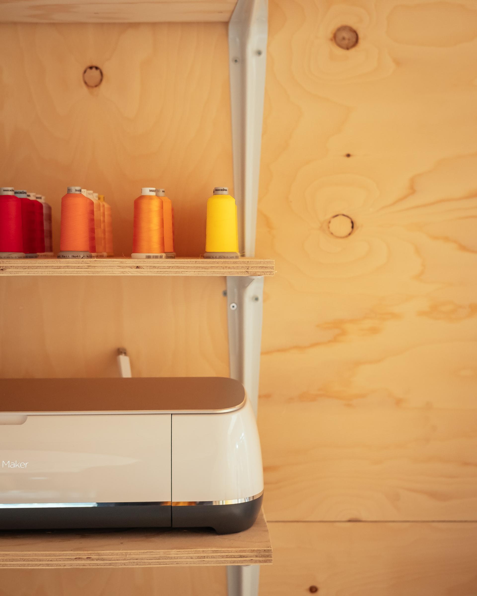 A row of colourful thread spools sits on a wooden shelf above a cutting machine, all set against a light wood panelled wall crafted from sustainably sourced materials with visible knots.
