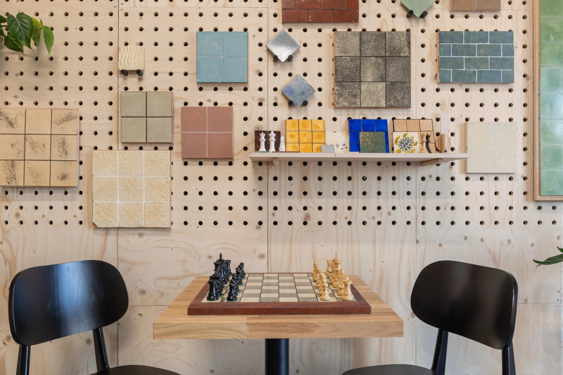 Two black chairs face a square table with a chessboard and pieces. The pegboard wall displays colourful tile samples from the Dekiln Showroom, with some plants partially visible on the sides.