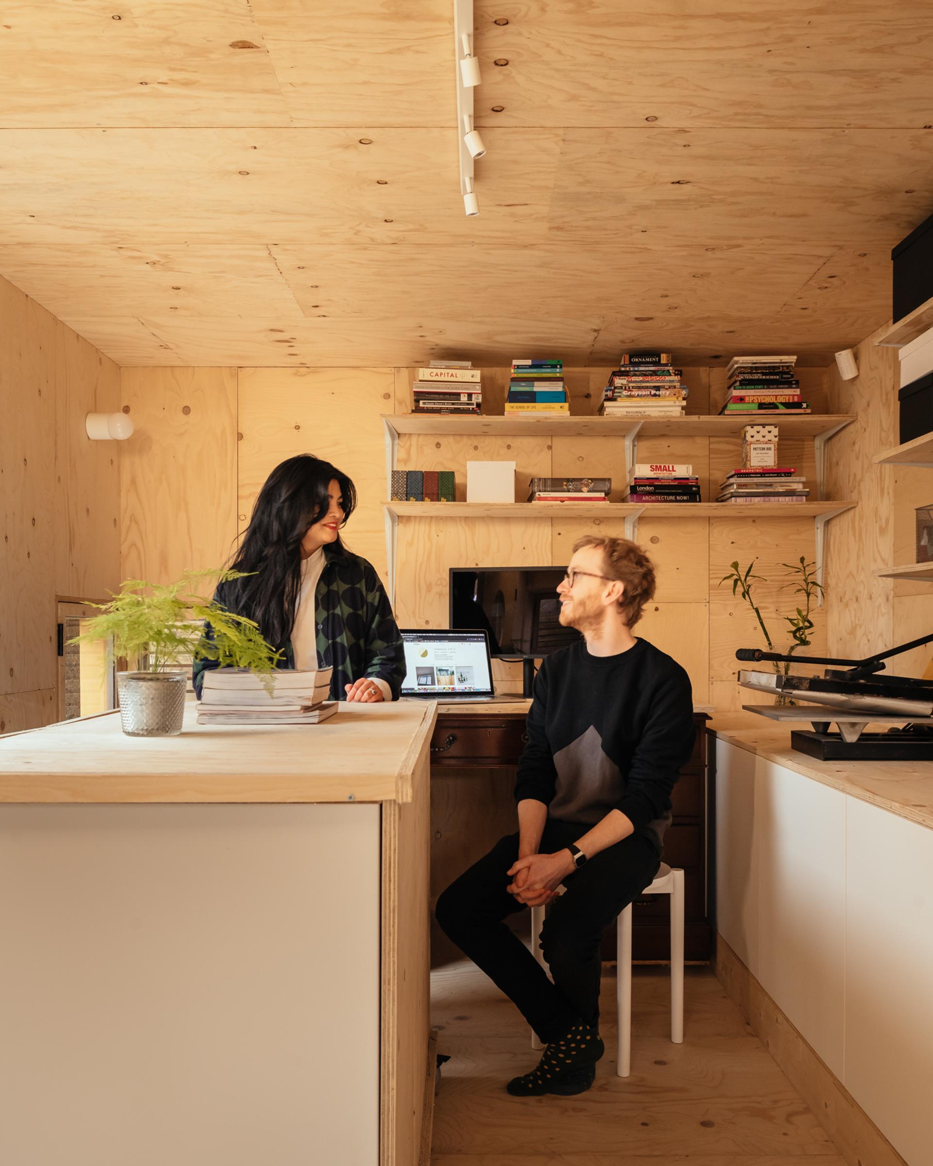 A woman stands behind a wooden counter, smiling at a seated man in a cosy, minimalist room with birch plywood walls, shelves of books, plants, and a laptop on a small desk in the background.