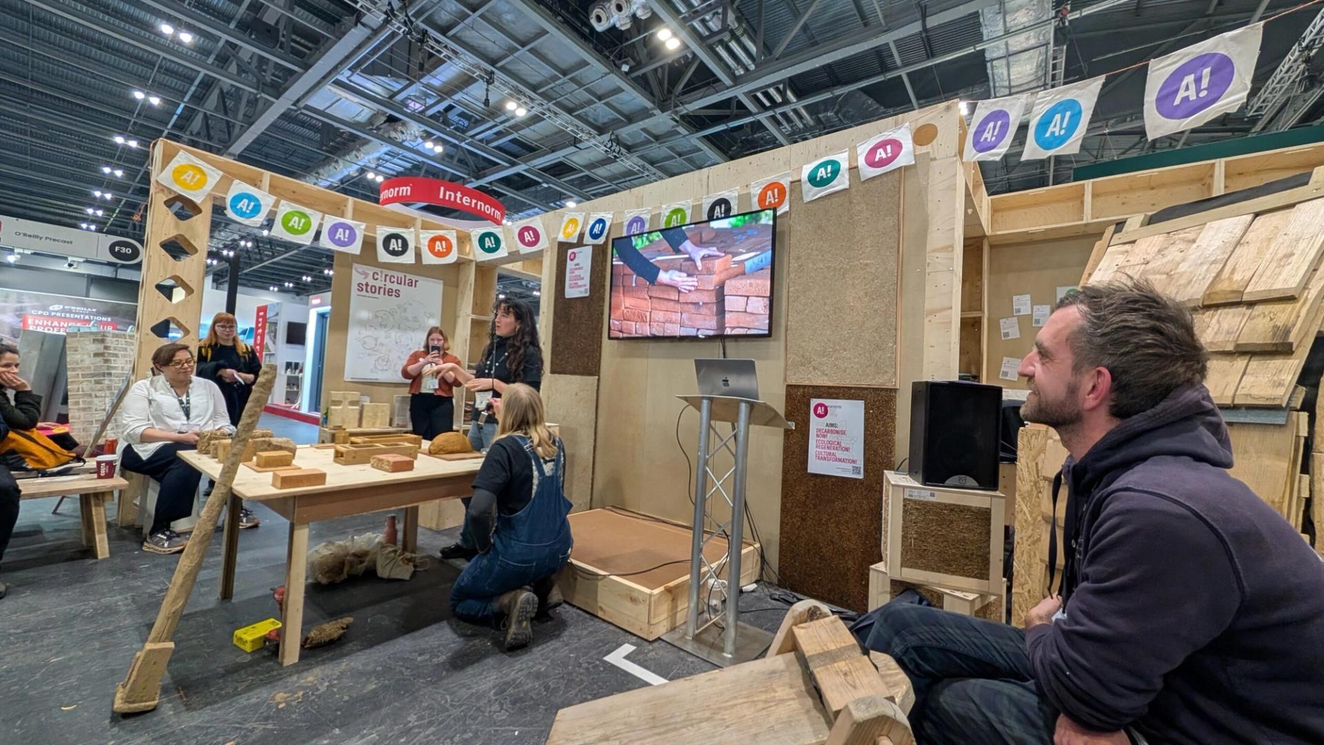 People sit and listen to a presentation about sustainable, eco-friendly building at an indoor exhibition stand, surrounded by wood and straw displays, with a monitor showing a brick wall and colourful banners overhead.