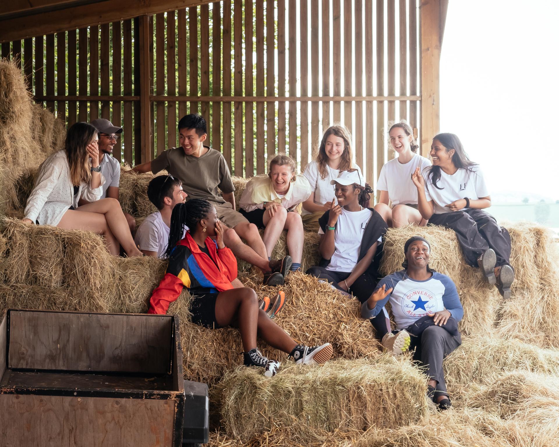 A diverse group of young people are sitting and laughing together on hay bales inside a birch timber construction barn, enjoying each other's company in a relaxed, casual setting.