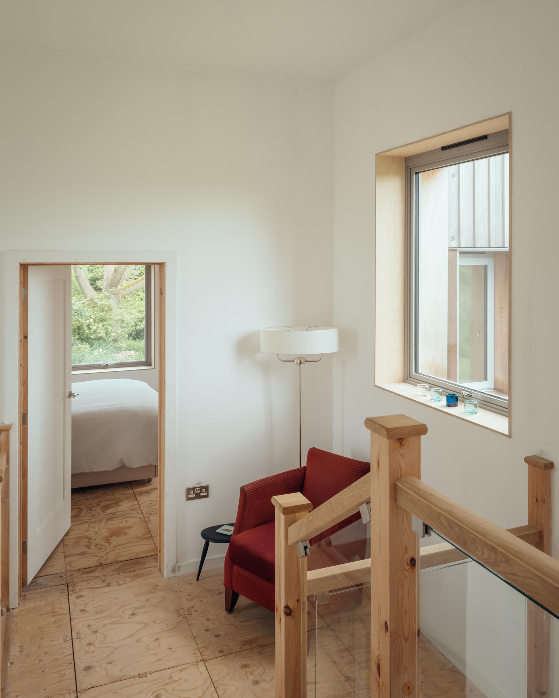 A bright hallway with wooden floors and stair railing built using the U-Build system, featuring a red armchair, small round black table, white floor lamp, and large window overlooking greenery. An open door leads to a bedroom.