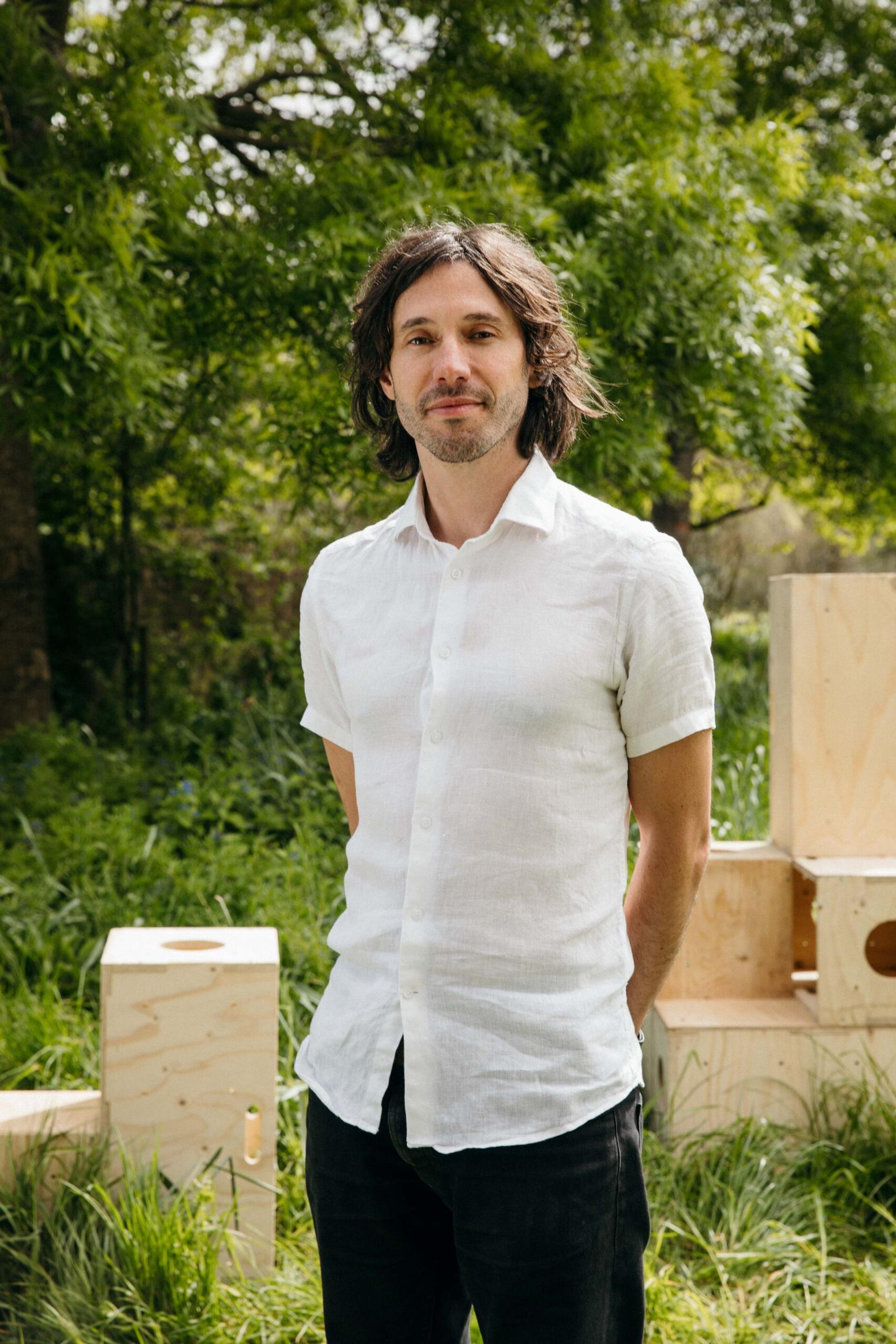 A man with medium-length dark hair, wearing a short-sleeved white shirt and black trousers, stands outdoors in a grassy area surrounded by leafy trees and sustainable timber construction featuring spruce wooden boxes.