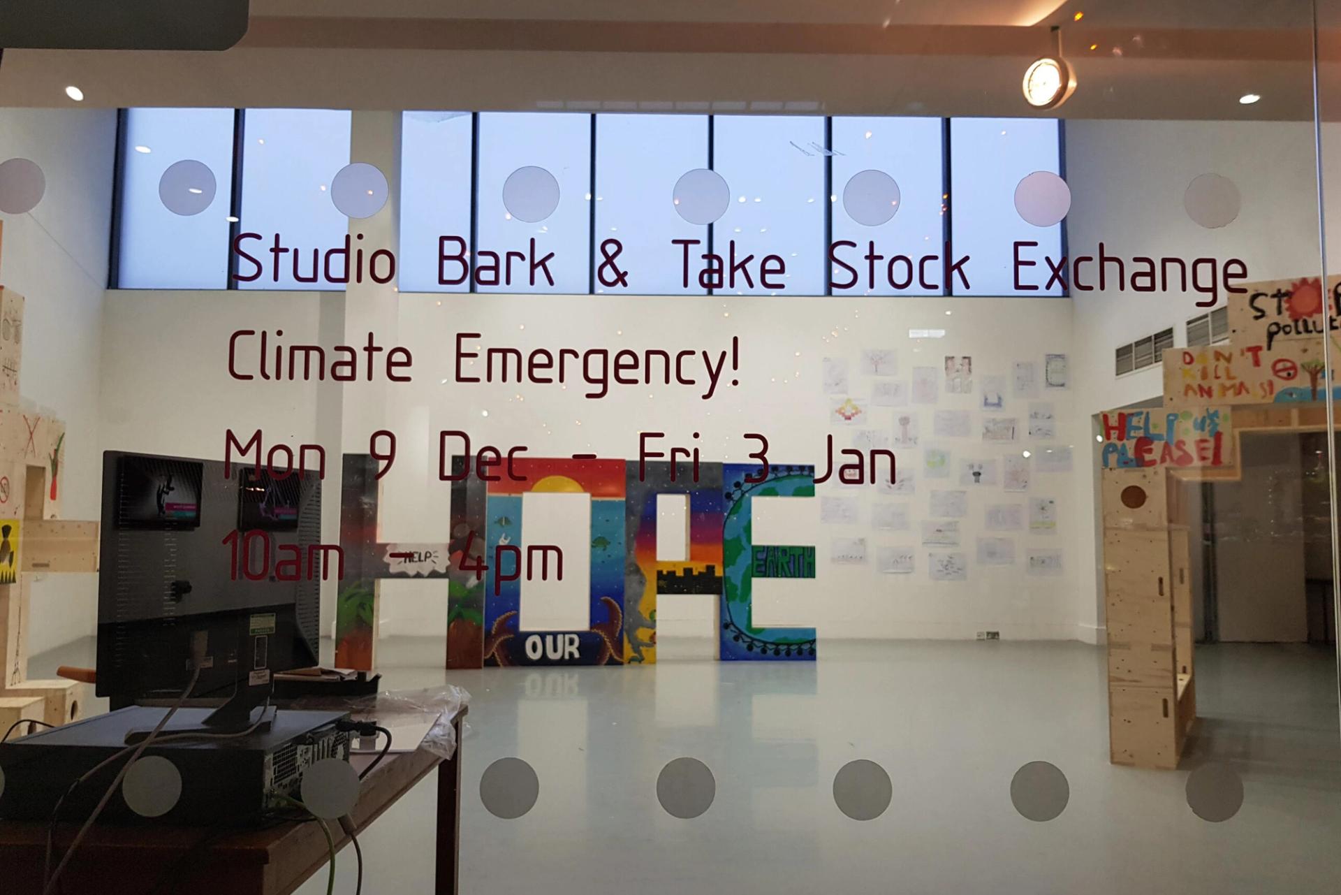 Glass door with red text advertising Studio Bark & Take Stock Exchange Climate Emergency! Mon 9 Dec – Fri 3 Jan, 10am – 4pm. Colourful artwork promoting upcycle and circular economy ideas is visible inside the bright room.