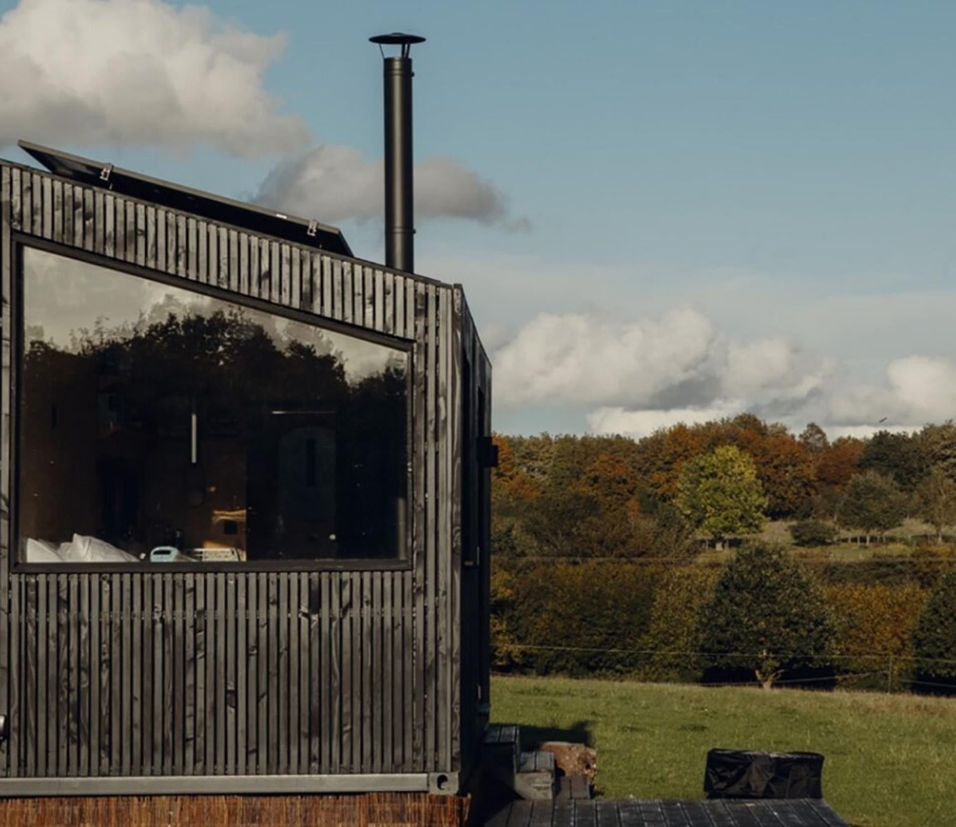 A modern, dark wood modular tiny house with large windows sits in a grassy field, surrounded by trees and under a blue sky with clouds.