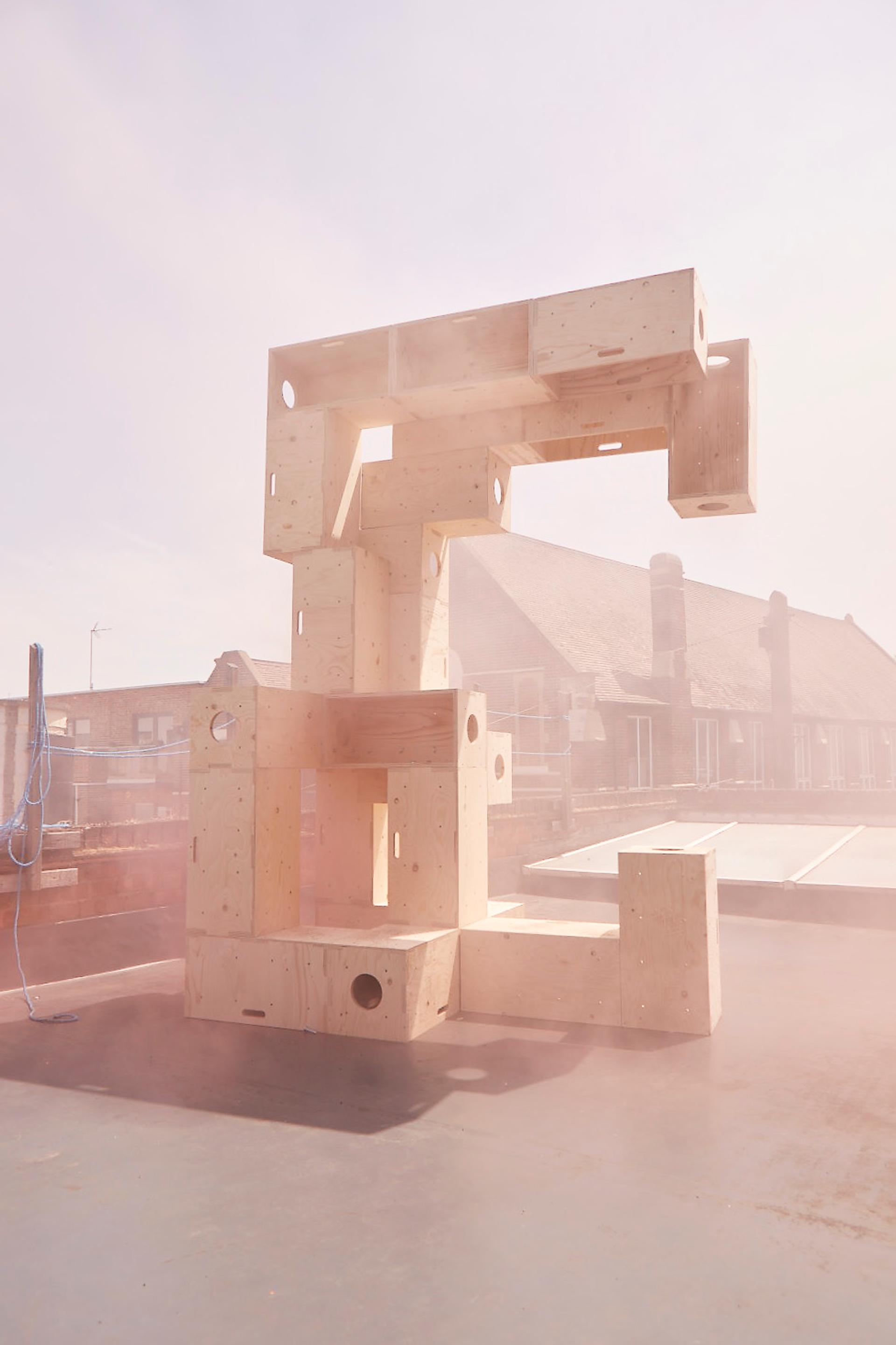 A tall sculpture made of large, modular plywood blocks stacked in an abstract, geometric shape stands outdoors on a rooftop, enveloped in a soft haze with buildings visible in the background.
