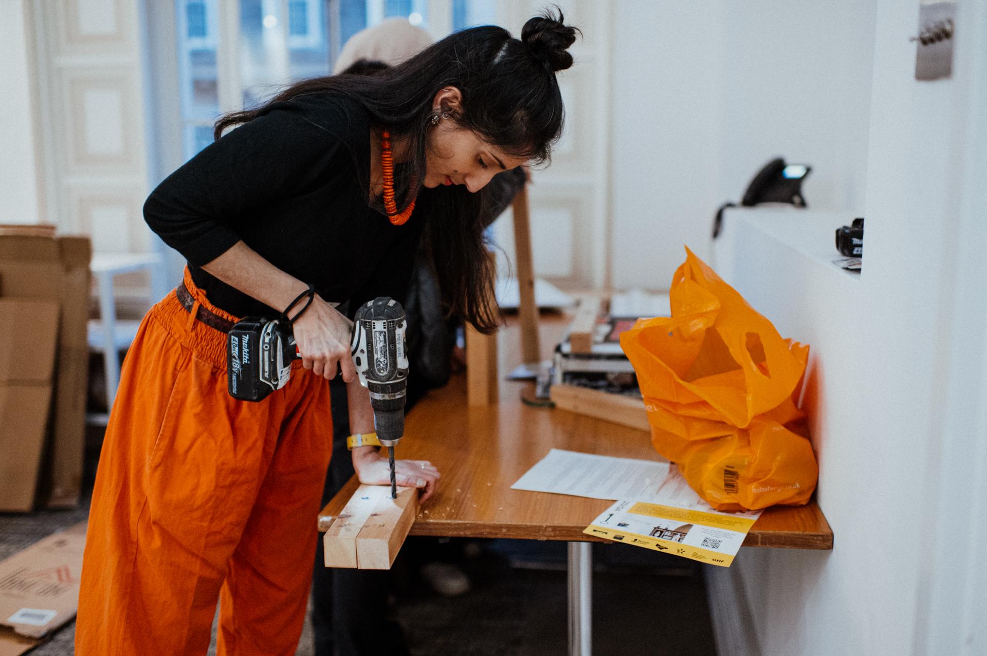 A woman in orange trousers uses a power drill on a wooden plank placed on a table, contributing to a youth architecture design community build project. Papers, an orange bag, and tools are scattered across the table in the well-lit room, suggesting an engaging hands-on environment focused on making and building creative structures.