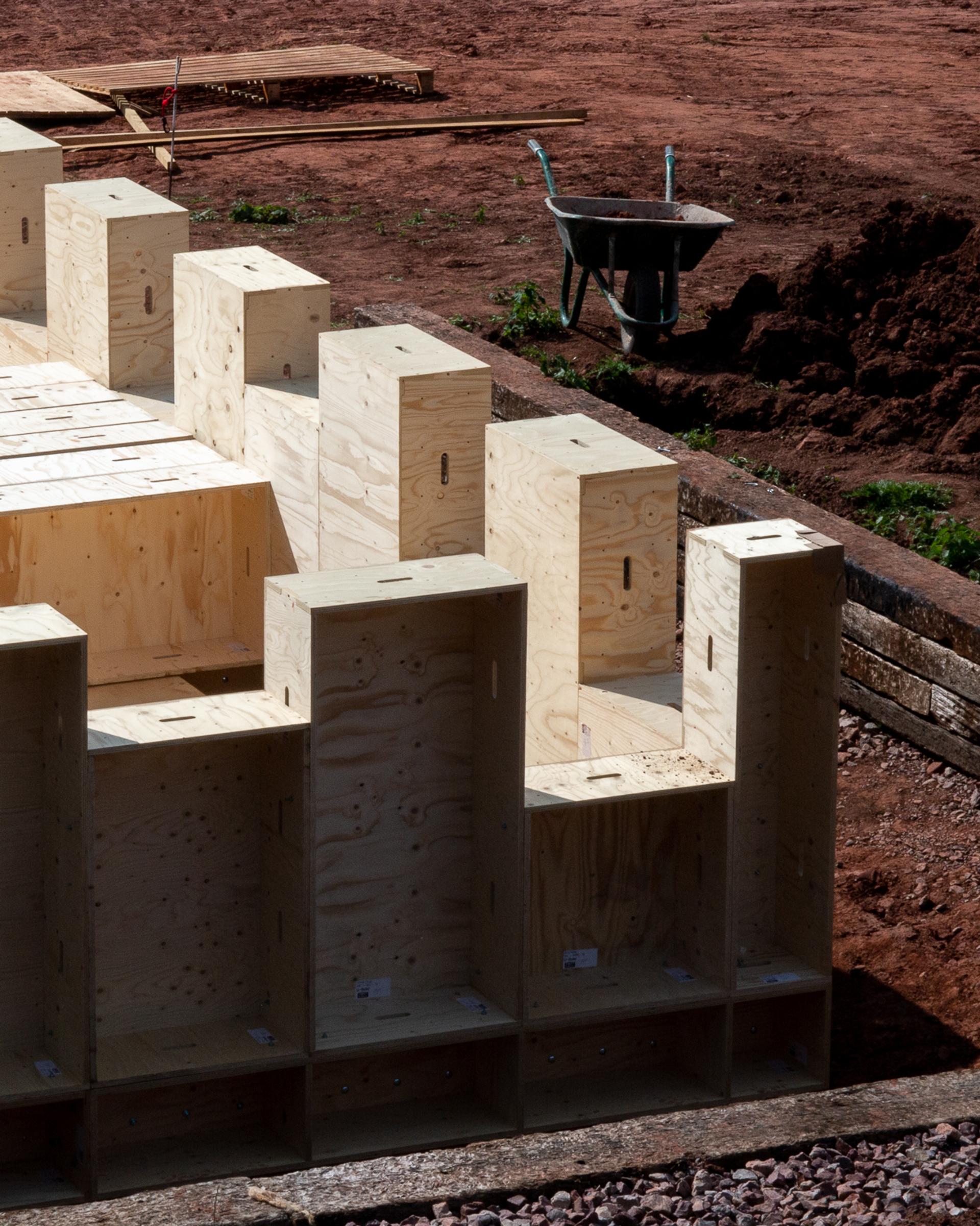 Several unfinished wooden cabinets or boxes, crafted from sustainably sourced timber, are arranged outdoors on reddish soil near a building site, with a wheelbarrow and wooden planks visible in the background.