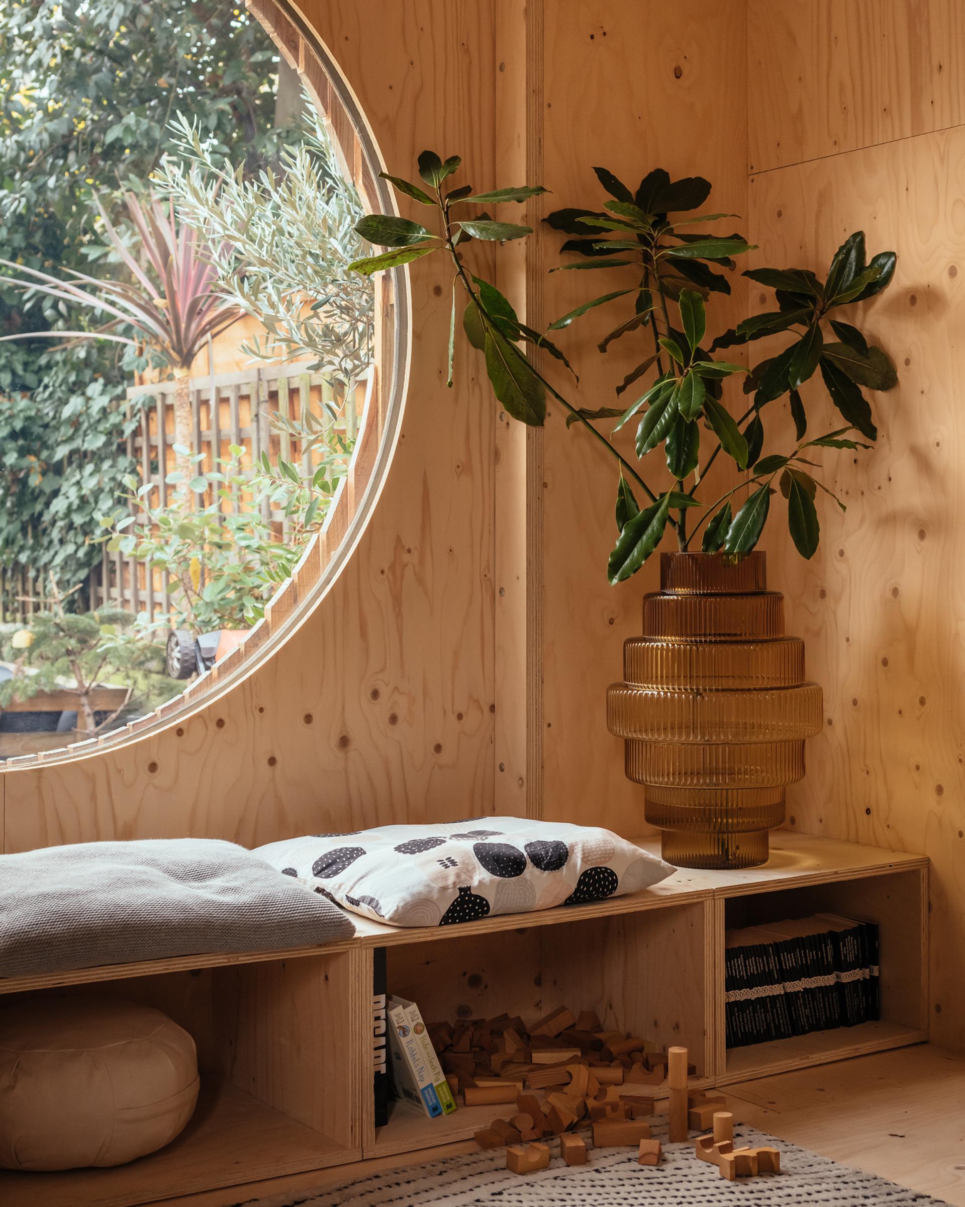 A cosy wooden nook crafted from sustainable birch, featuring a large round window, a cushion, decorative pillow, potted plant in a textured vase, wooden blocks, and a relaxing view of the lush garden outside.